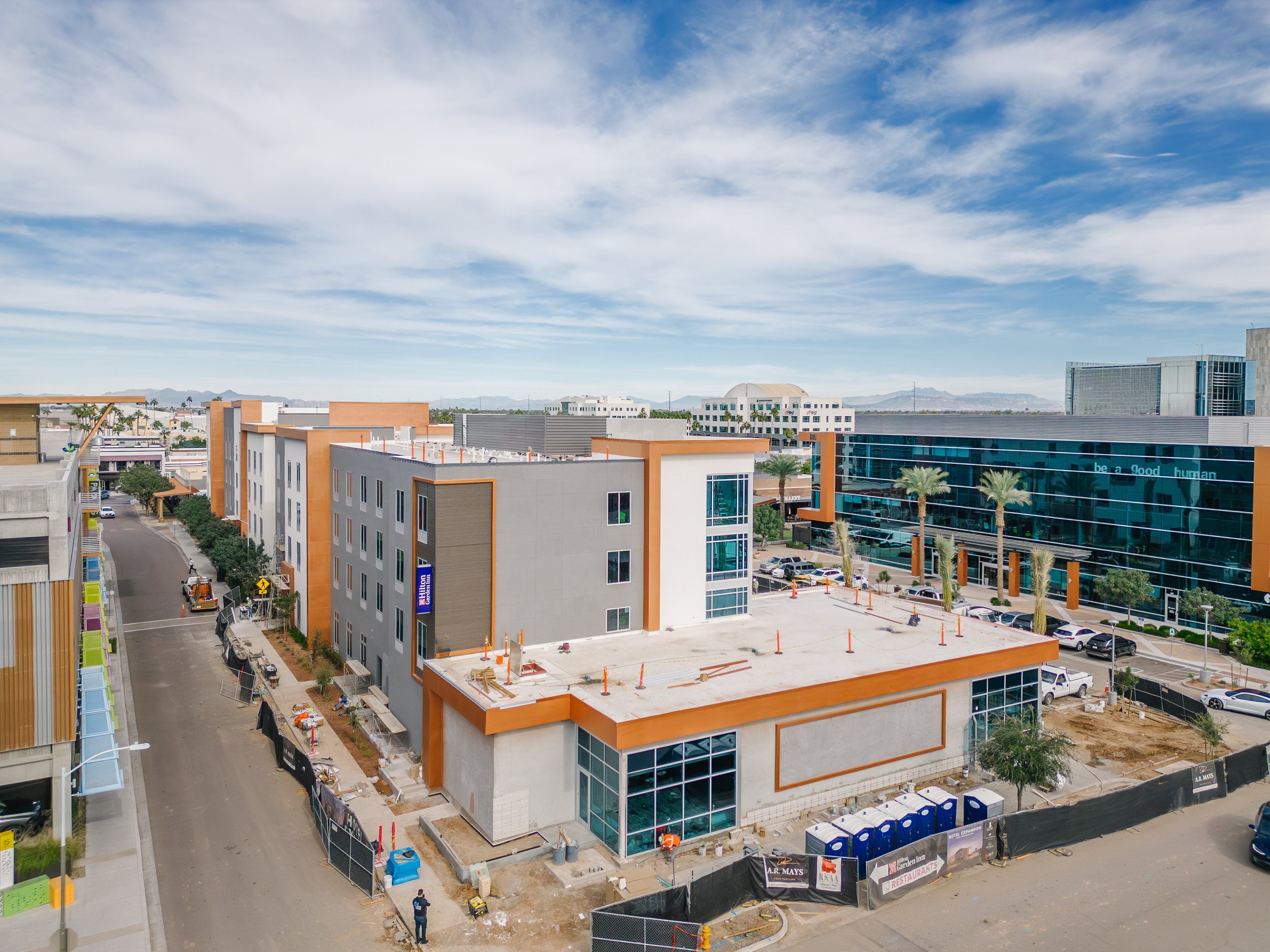 Construction site with a multi-story building under development, surrounded by parked cars and construction materials, in an urban area with modern buildings and blue sky.