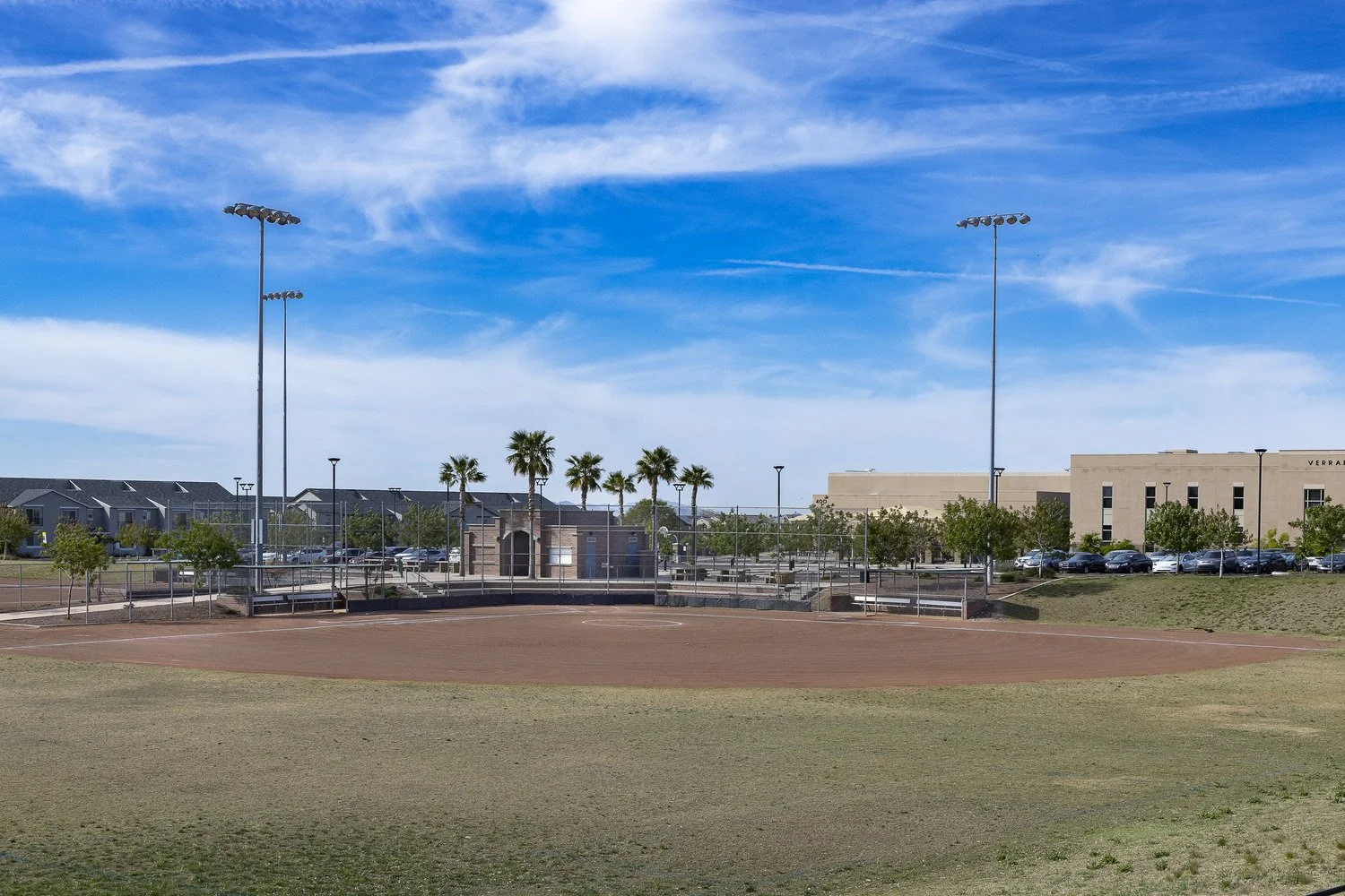 Empty baseball field with dirt infield and grass outfield, surrounded by chain-link fence, with a parking lot and buildings in the background, under a blue sky with scattered clouds.