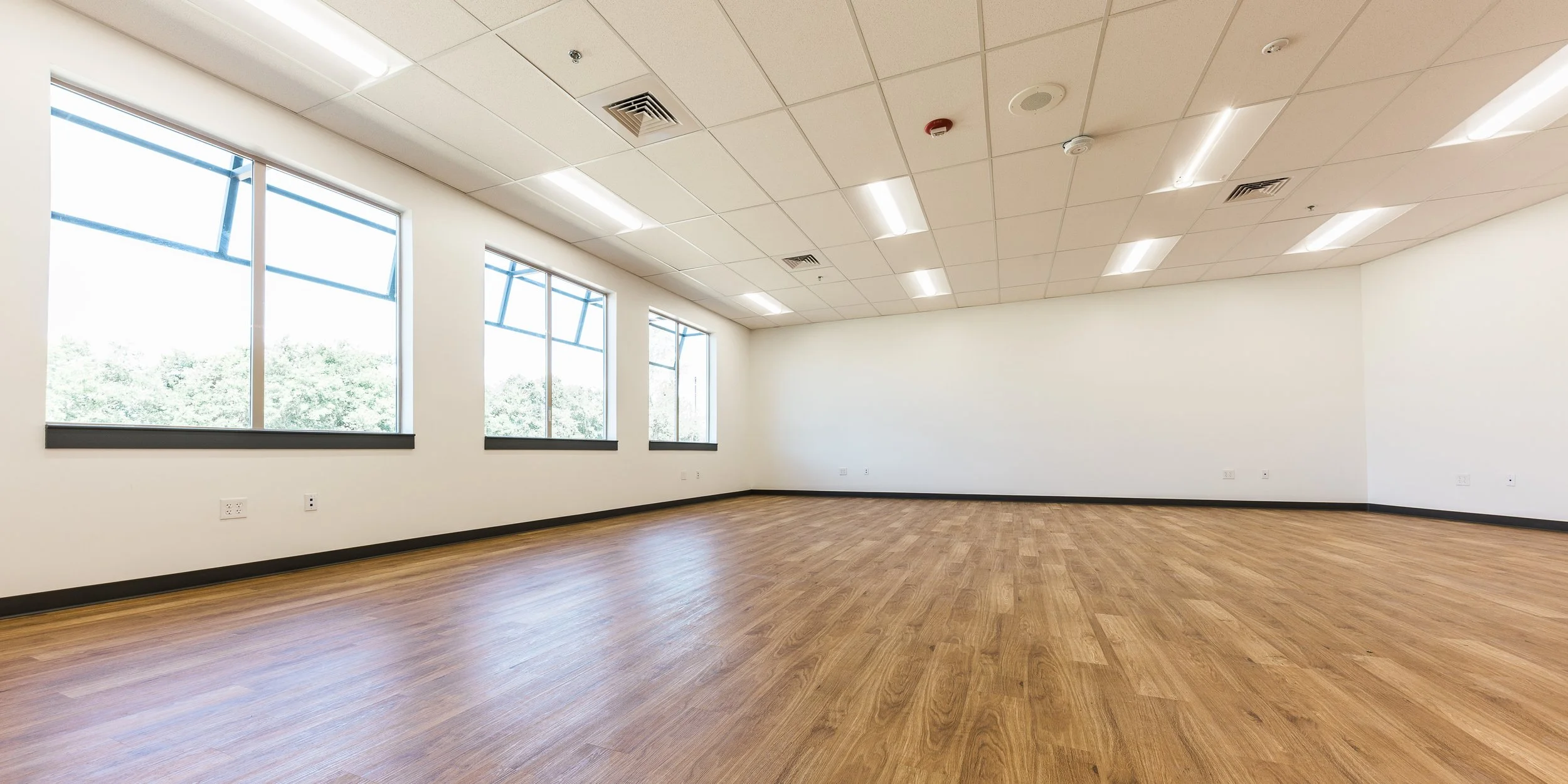 Empty room with large windows, white walls, wooden flooring, and ceiling lights.