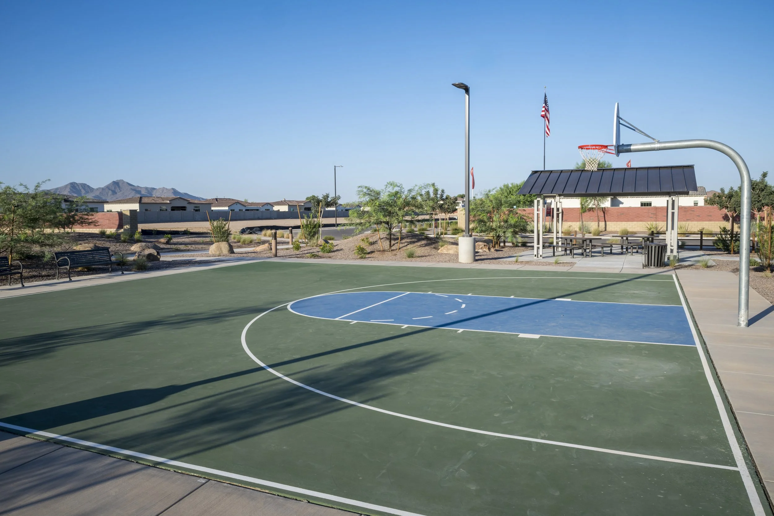 Empty outdoor basketball court with a hoop, shaded seating area, flags, and a mountain in the background.