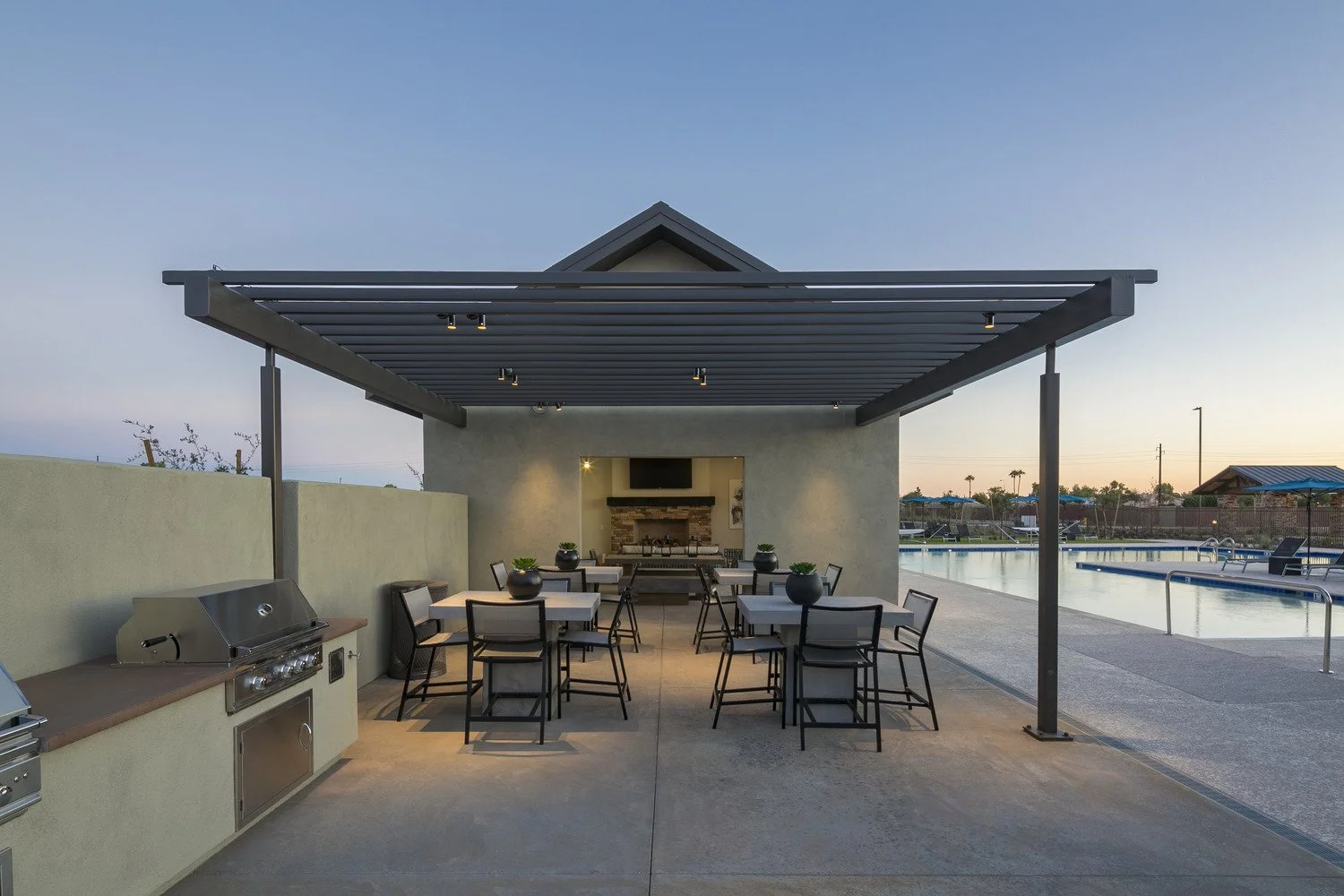 Outdoor poolside lounge area with a covered patio, tables, chairs, a grill, and a view of the swimming pool at sunset.