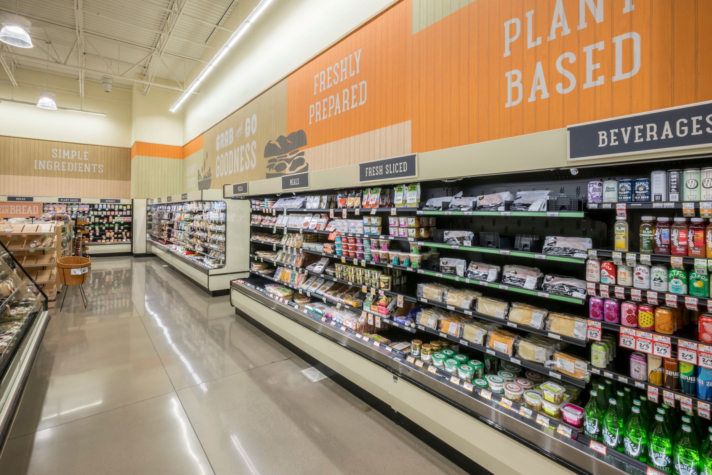 A grocery store aisle with shelves stocked with cheese, dairy, and plant-based food products, and signs indicating sections for meals, fresh sliced items, beverages, and simple ingredients.