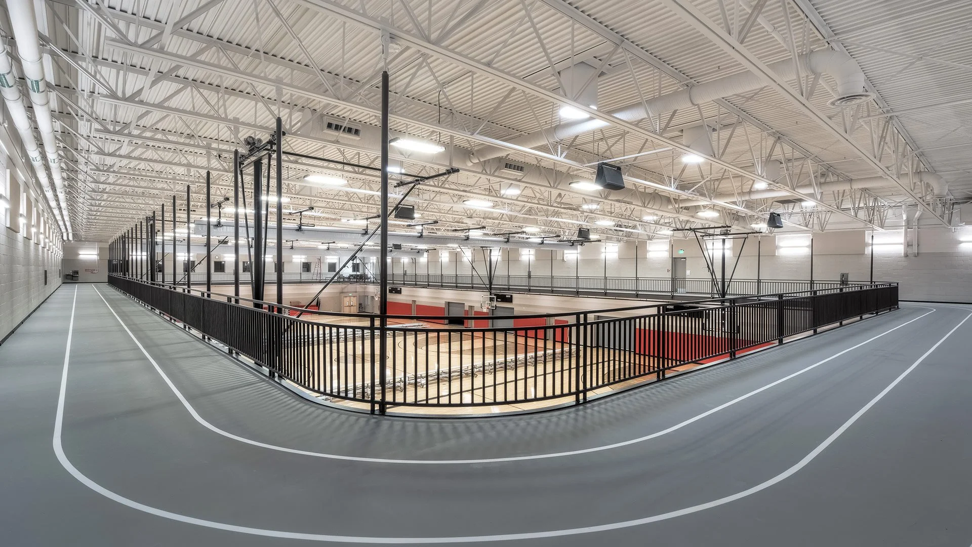 Indoor sports gymnasium with a running track surrounding a basketball court. The gym has high ceilings with exposed white metal beams and bright lighting.