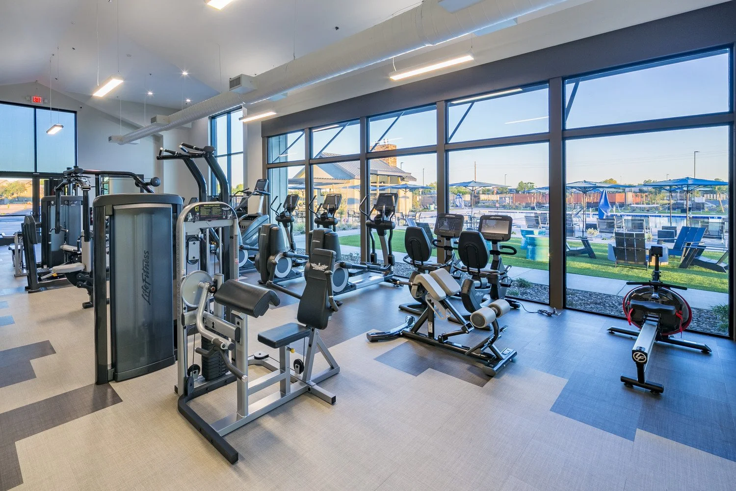 Indoor gym with various exercise machines, including ellipticals, stationary bikes, and strength training equipment, with large windows showing outdoor seating and water view.