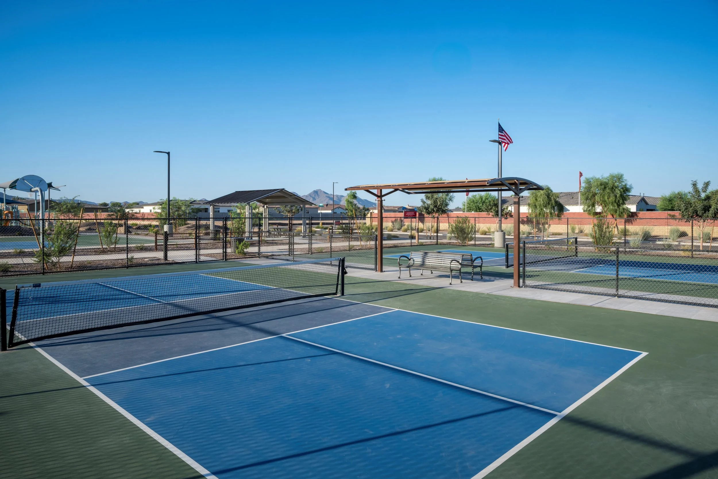 Empty outdoor tennis courts with blue and green surfaces, surrounded by a black chain-link fence, under a clear blue sky. There are benches and shade structures nearby, with an American flag flying on a pole.
