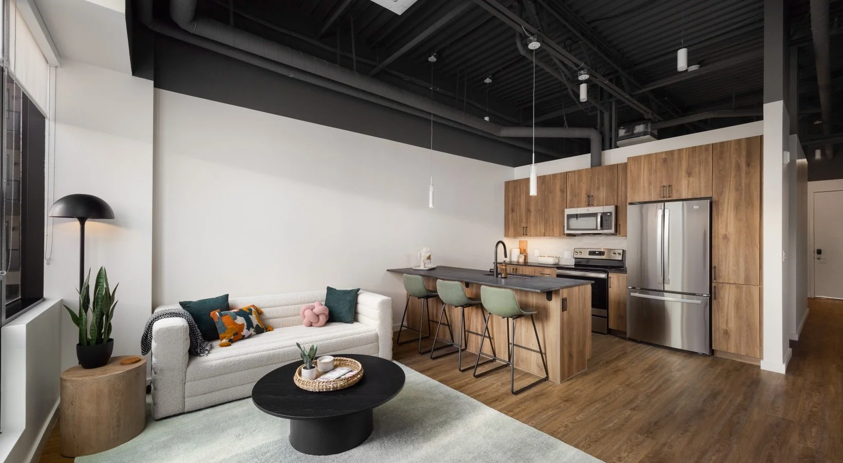 Open-concept living room and kitchen with modern decor, including a white sofa with colorful pillows, a black coffee table, a wooden side table with a plant, and kitchen featuring wooden cabinetry, stainless steel appliances, and a breakfast bar with