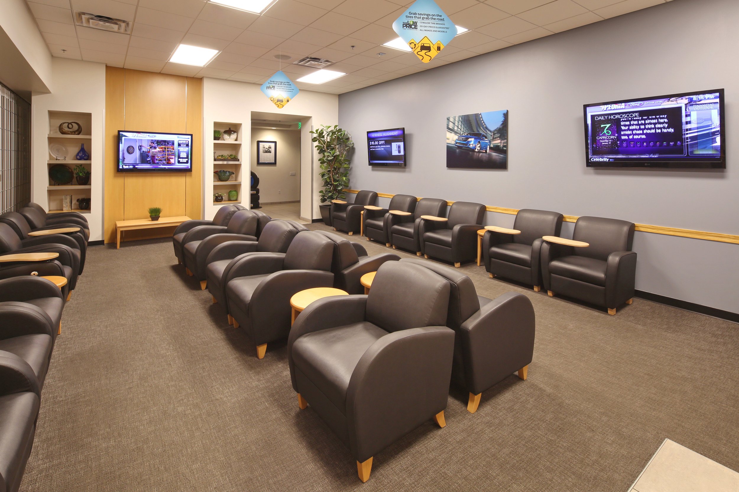 Empty waiting room with rows of black leather chairs, small wooden side tables, and wall-mounted TV screens displaying information and advertisements. There are decorative shelves with vases and plants, a large potted plant near the entrance, and cei