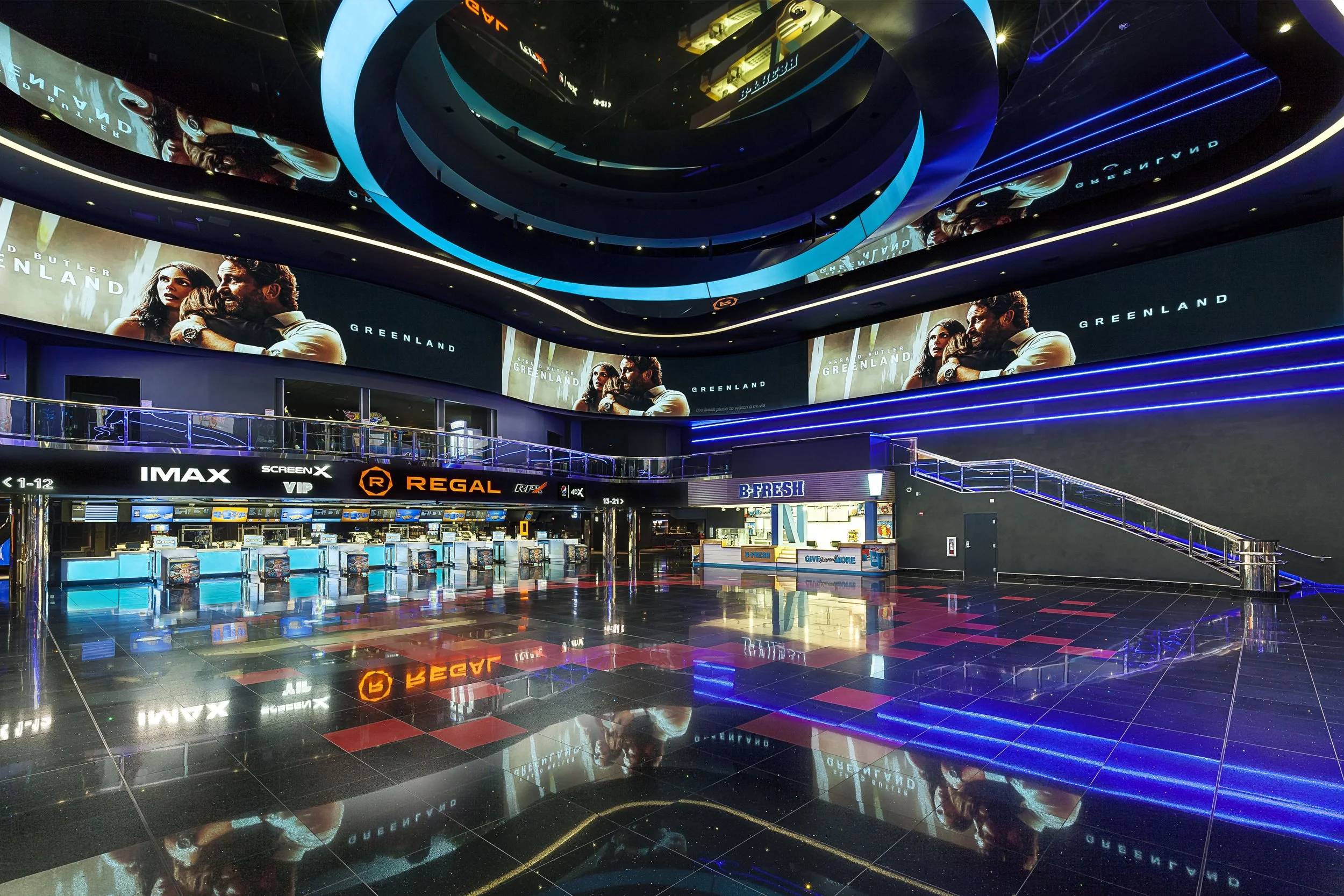 Inside a modern movie theater lobby with digital screens, ticket kiosks, and a staircase illuminated with blue neon lights.