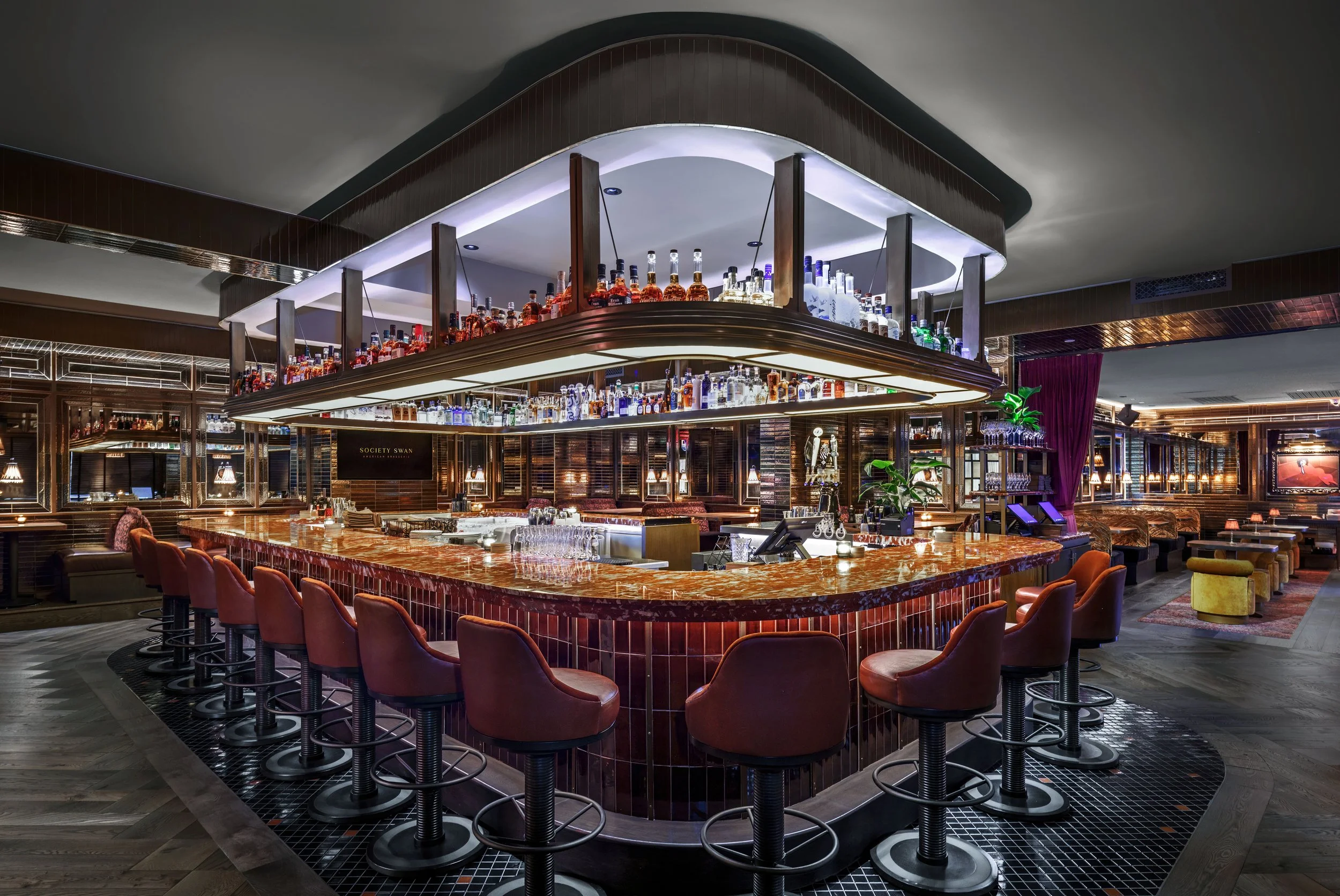 Interior of a stylish, upscale bar with a circular marble countertop, high-backed bar stools, and a suspended liquor display above. Warm lighting and modern decor create an inviting atmosphere.