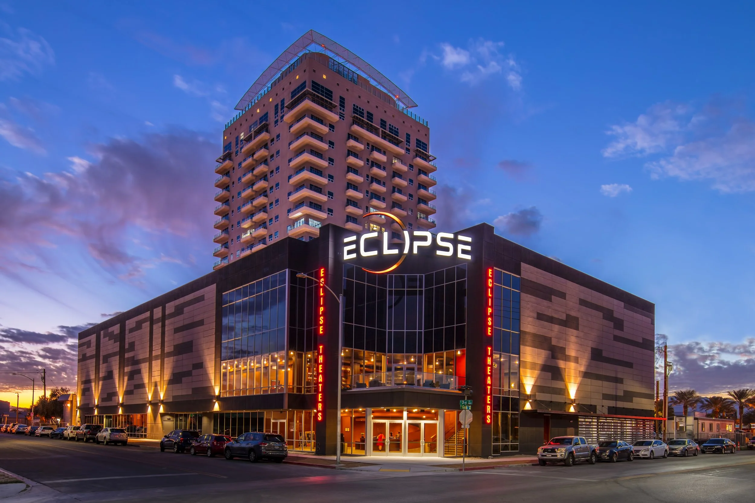 A modern building with glass windows and a sign that reads 'ECLIPSE' at dusk, with a tall residential tower in the background, parked cars along the street, and a colorful sky.
