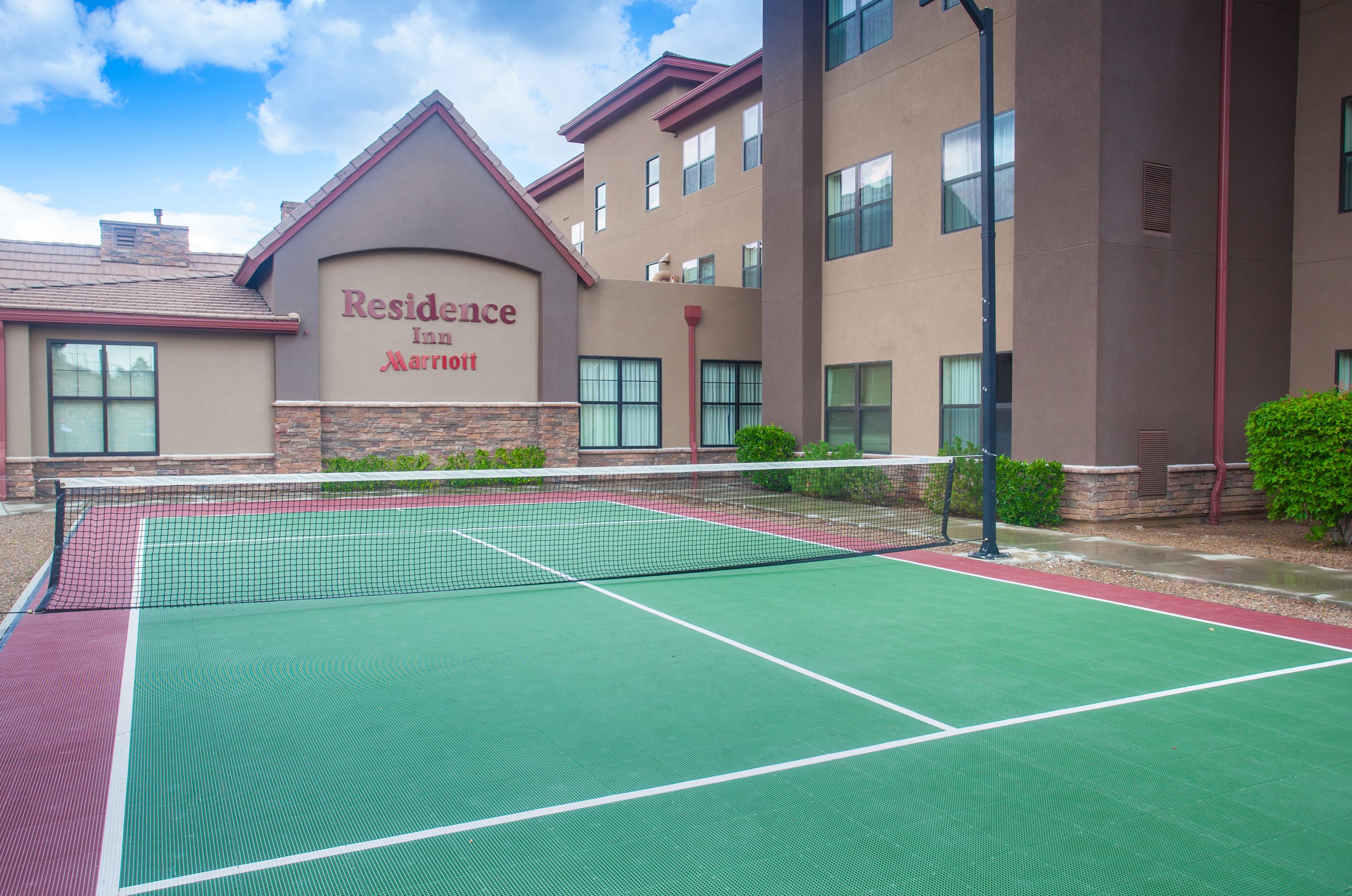Outdoor tennis court with green surface and white lines in front of a multi-story residence inn/marriott building.