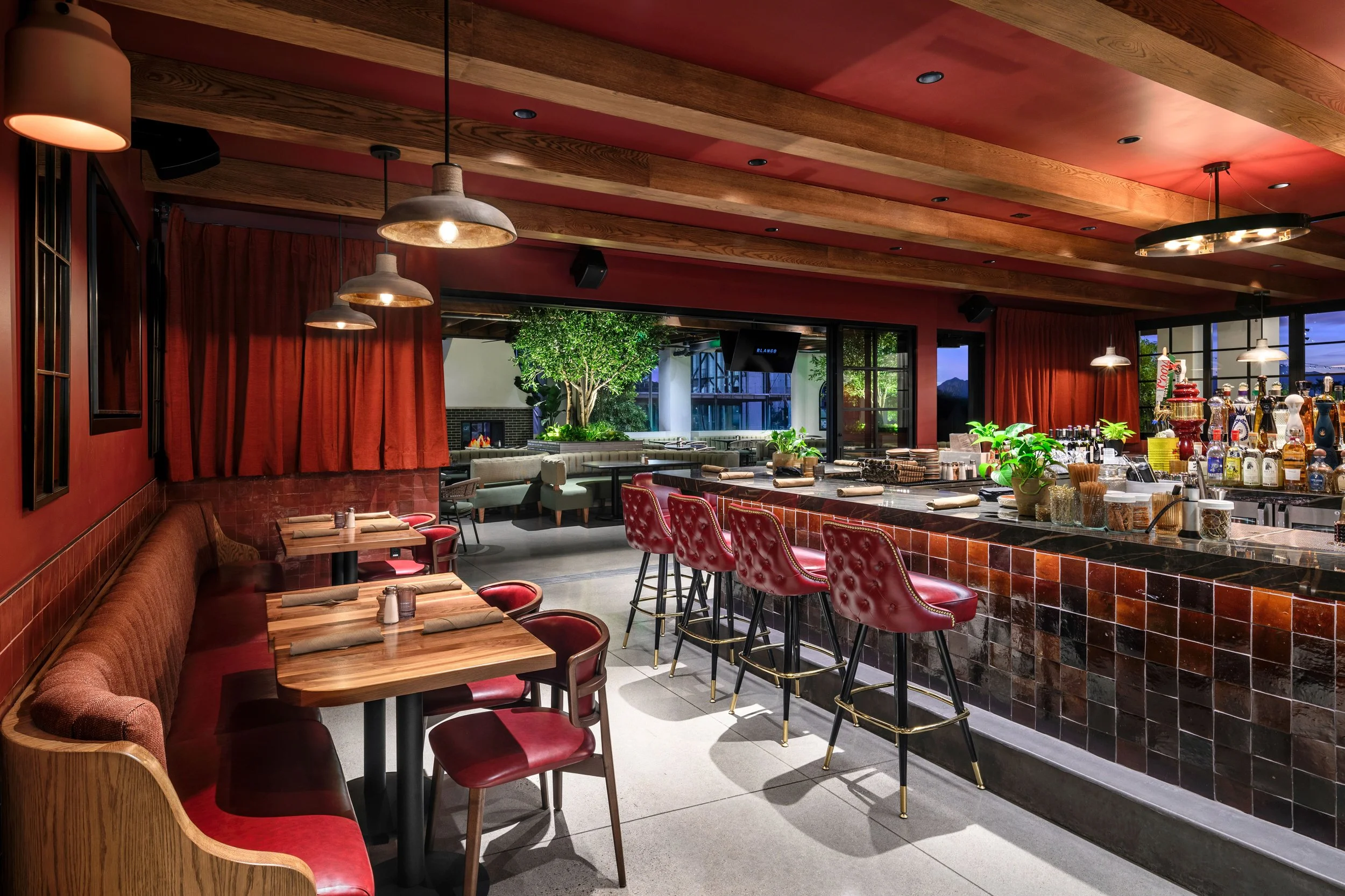 Interior of a restaurant or bar with red walls, wooden ceiling, and warm lighting. There are tables with red chairs, a marble bar counter with plants and bottles, and an outdoor patio area with greenery visible through large glass windows.