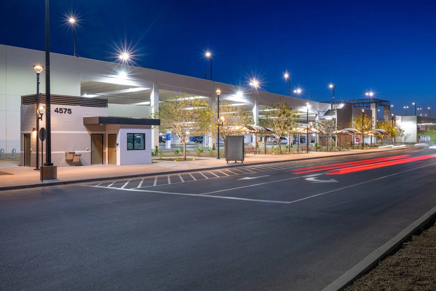 An illuminated parking area and a multi-story parking garage at night. The scene shows streetlights, trees, and a small building with the number 4575.