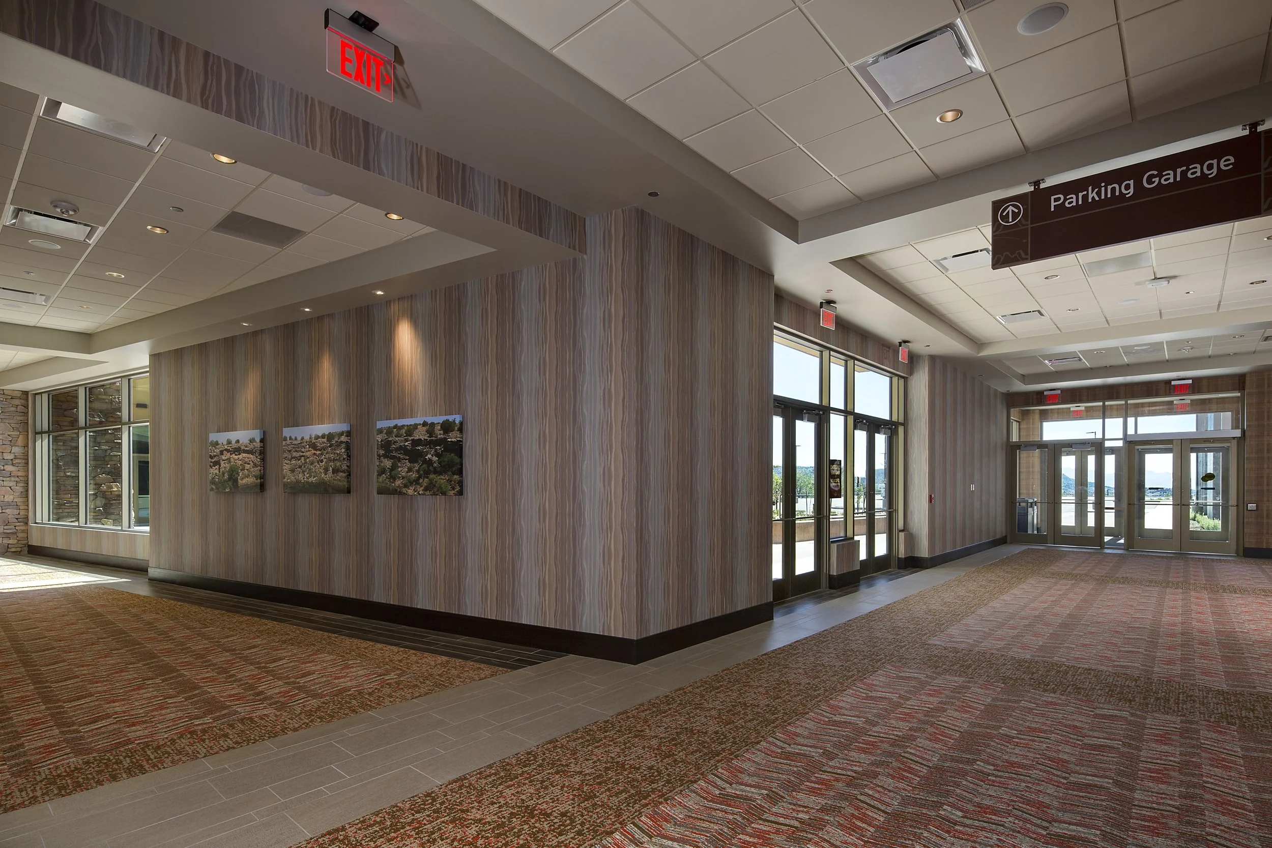 Empty indoor lobby area with glass doors leading outside, wooden wall with three landscape photographs, patterned carpet, ceiling lights, and red exit signs.