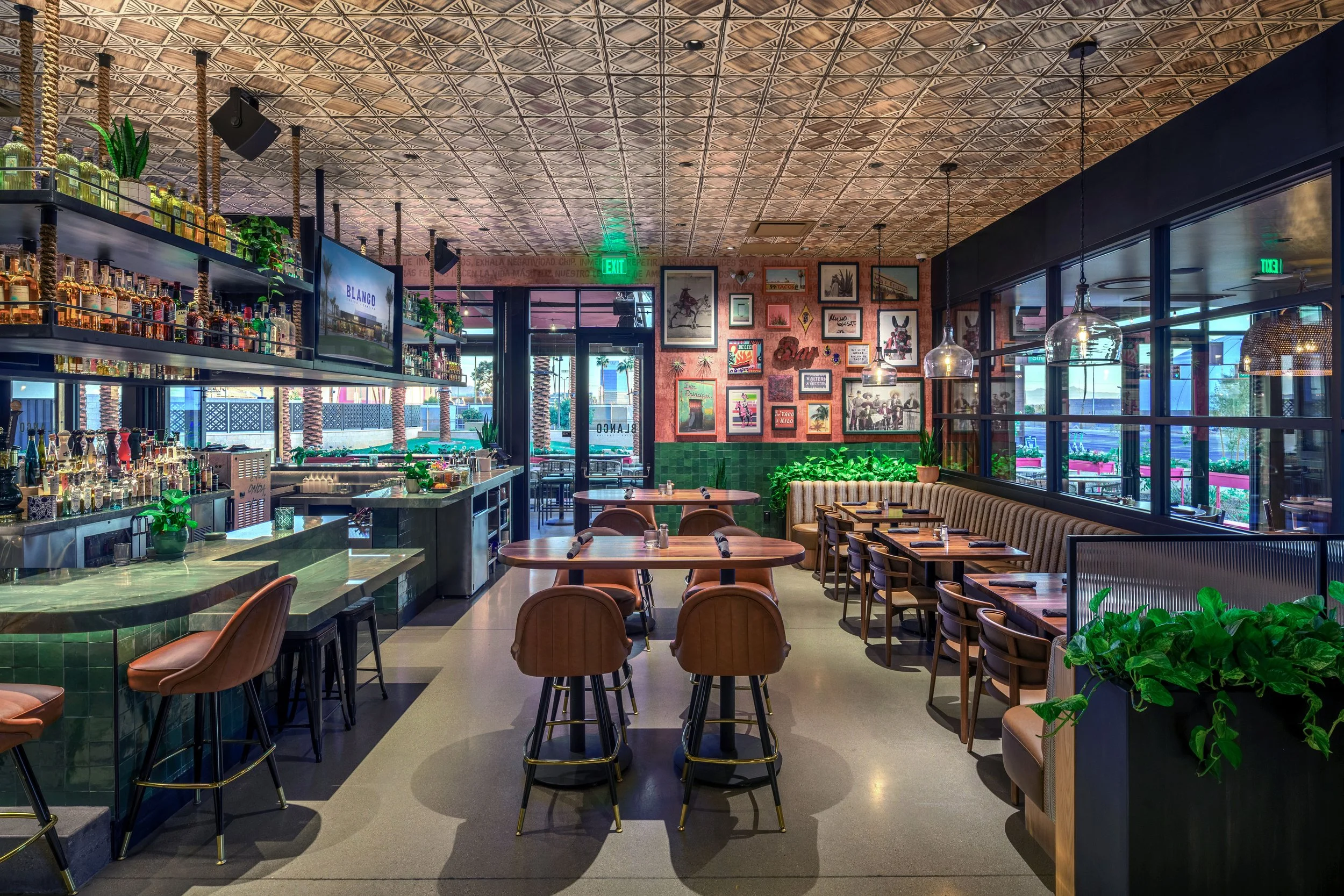 Interior of a modern restaurant or bar with a colorful, artistic decor, green tiled bar area, framed art on a pink wall, tables set with utensils, and large windows allowing natural light.