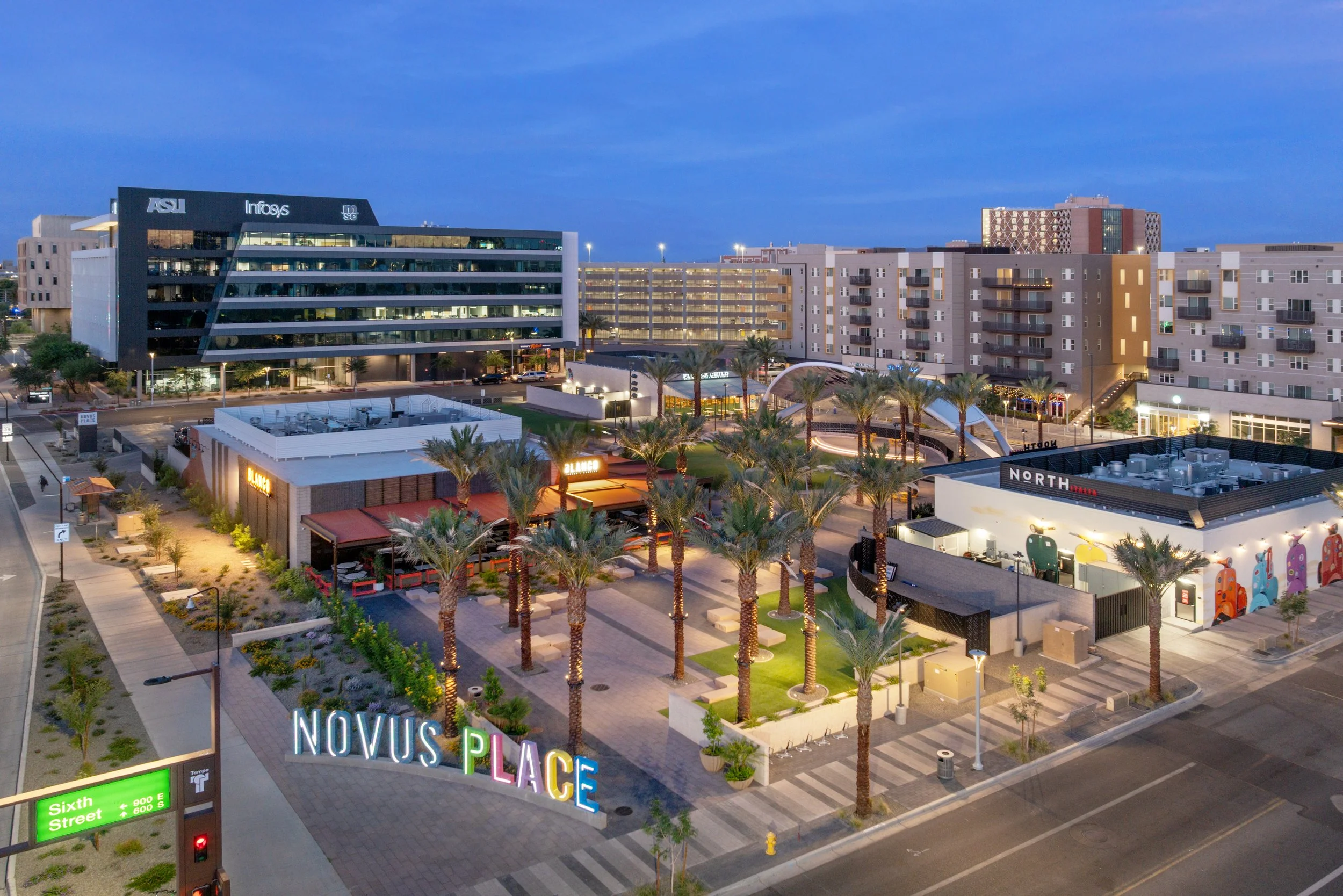 Aerial view of Novus Place shopping area at dusk featuring palm trees, outdoor seating, and modern buildings with signage, surrounded by streets and parking lots.