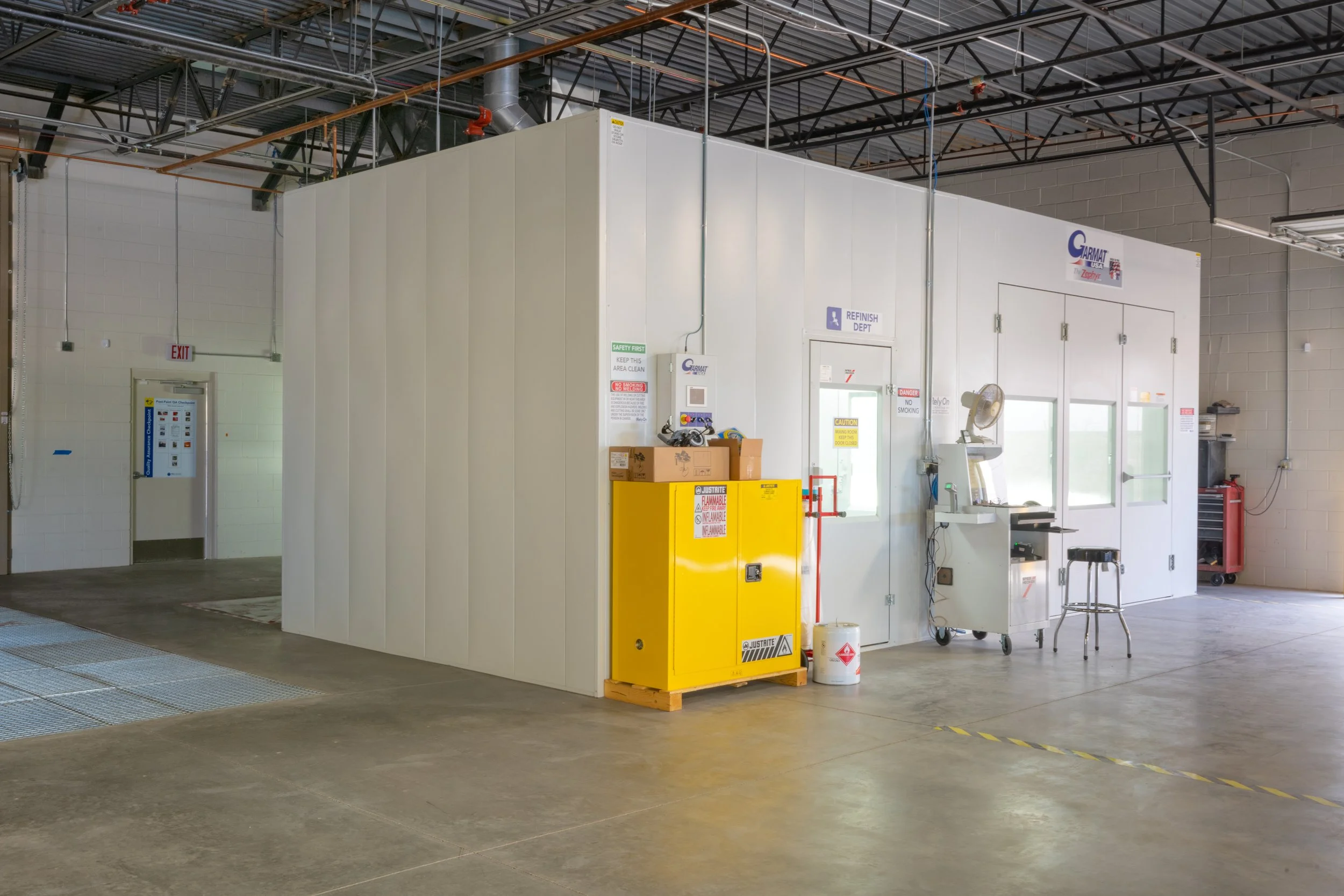 Industrial workspace with a white enclosed room, yellow safety cabinet, tools, and equipment on a plain concrete floor.