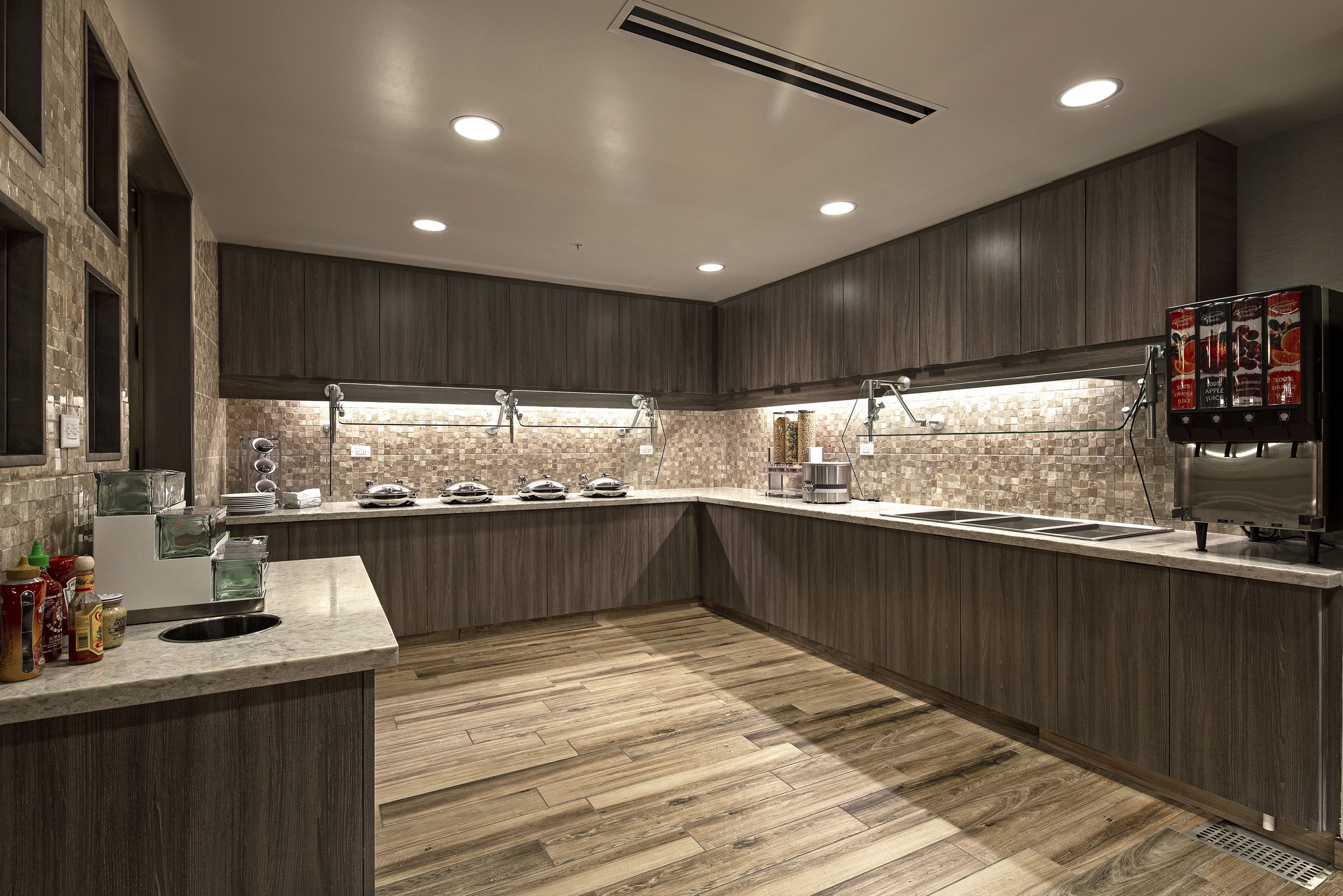 Empty buffet area in a hotel or restaurant kitchen with wood cabinets, a coffee station, and a drink dispenser.