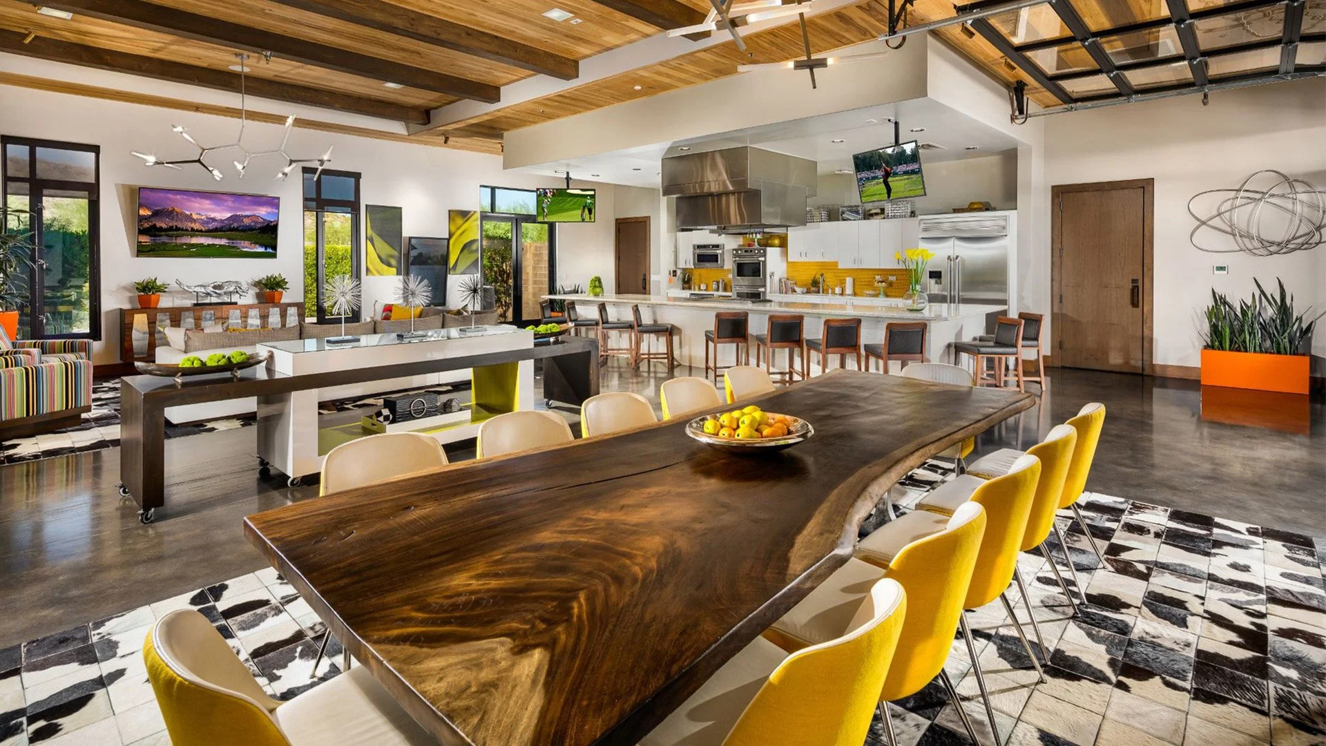 Open-concept modern living space with wooden ceiling beams, a kitchen with white cabinets and stainless steel appliances, a large wooden dining table with yellow chairs, multiple television screens, and contemporary artwork and decor.