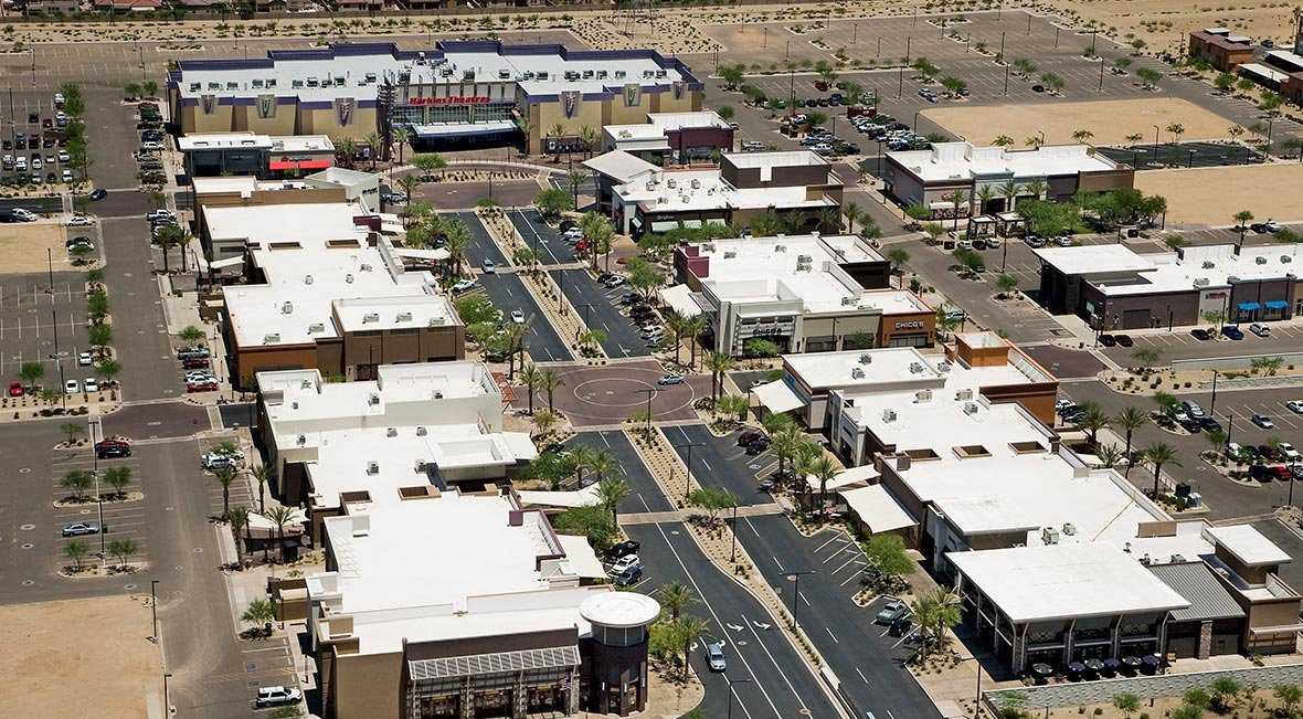 Aerial view of a modern shopping center with white rooftops, parking lots, and palm trees, located in a desert area.