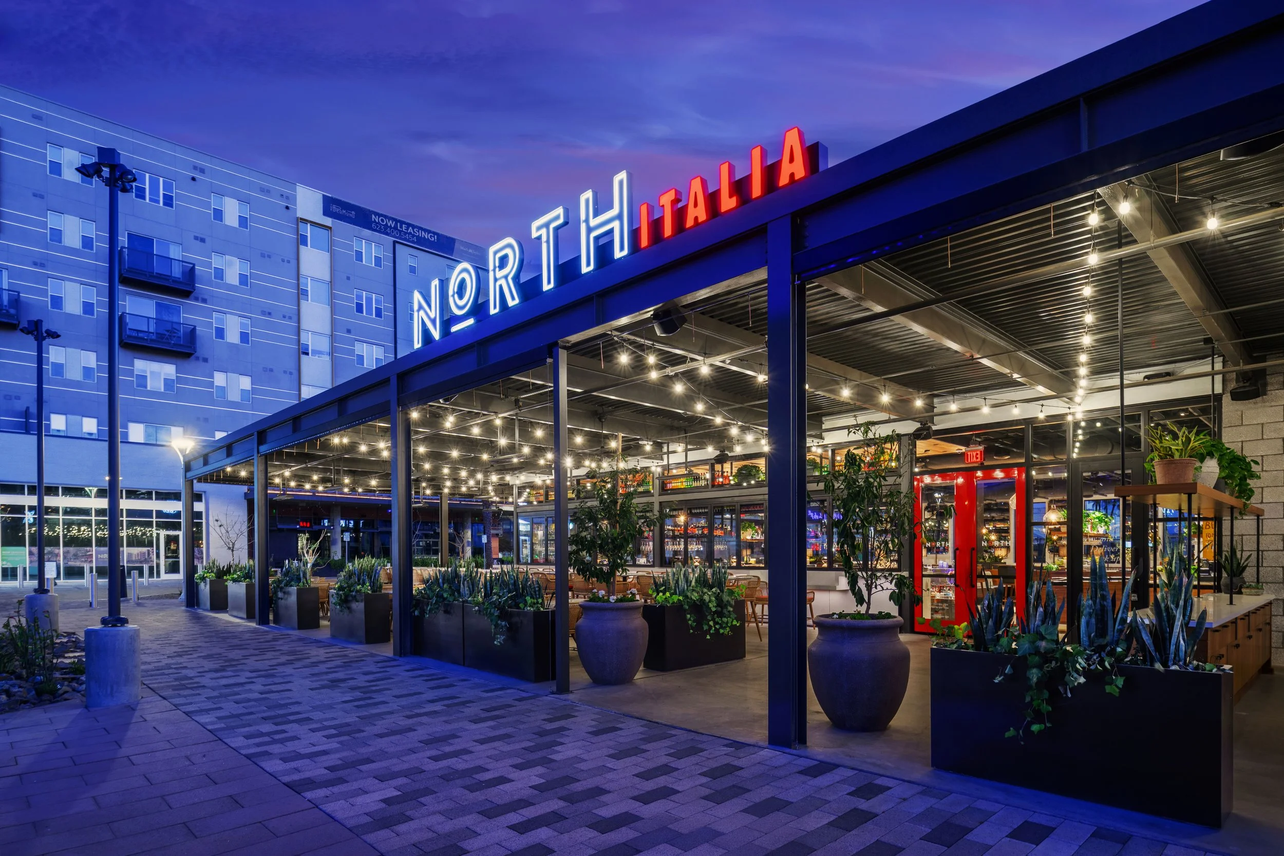 Night view of restaurant named North Italia with illuminated sign, outdoor patio with string lights, potted plants, and indoor seating visible through glass walls.