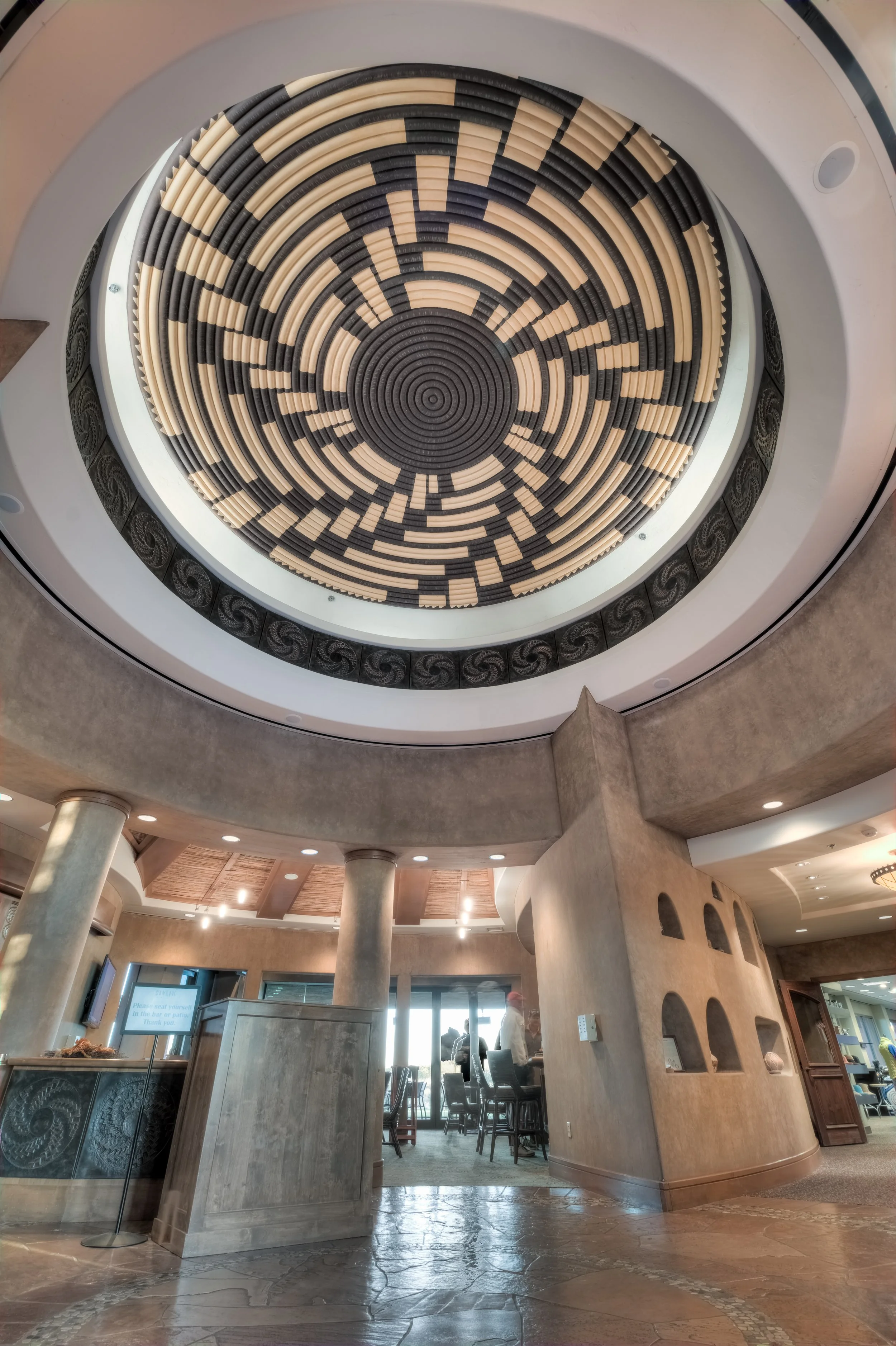 Interior view of a restaurant or cafe with a decorative ceiling featuring a circular pattern of black and beige tiles. The space has columns, textured walls, and a mix of tables and chairs, with some people visible near the windows.