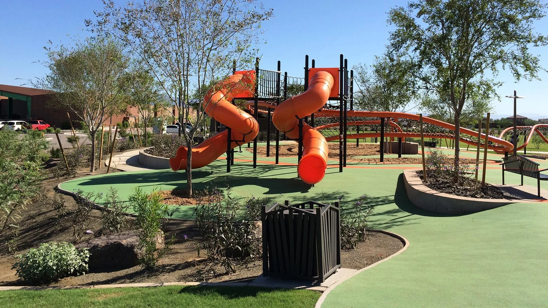 An empty playground with orange slides and a climbing structure, surrounded by trees, benches, and landscaped gardens on a sunny day.