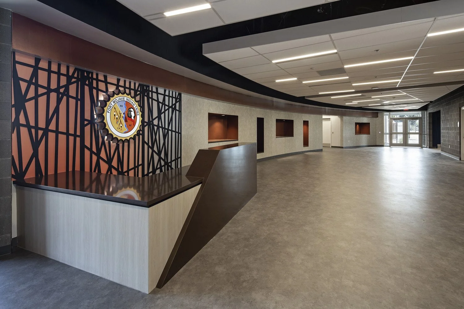 Empty lobby area of a government building with a curved reception desk and wall logo for Fort Mojave Indian Tribe, featuring modern design with recessed shelves and glass doors at the entrance.