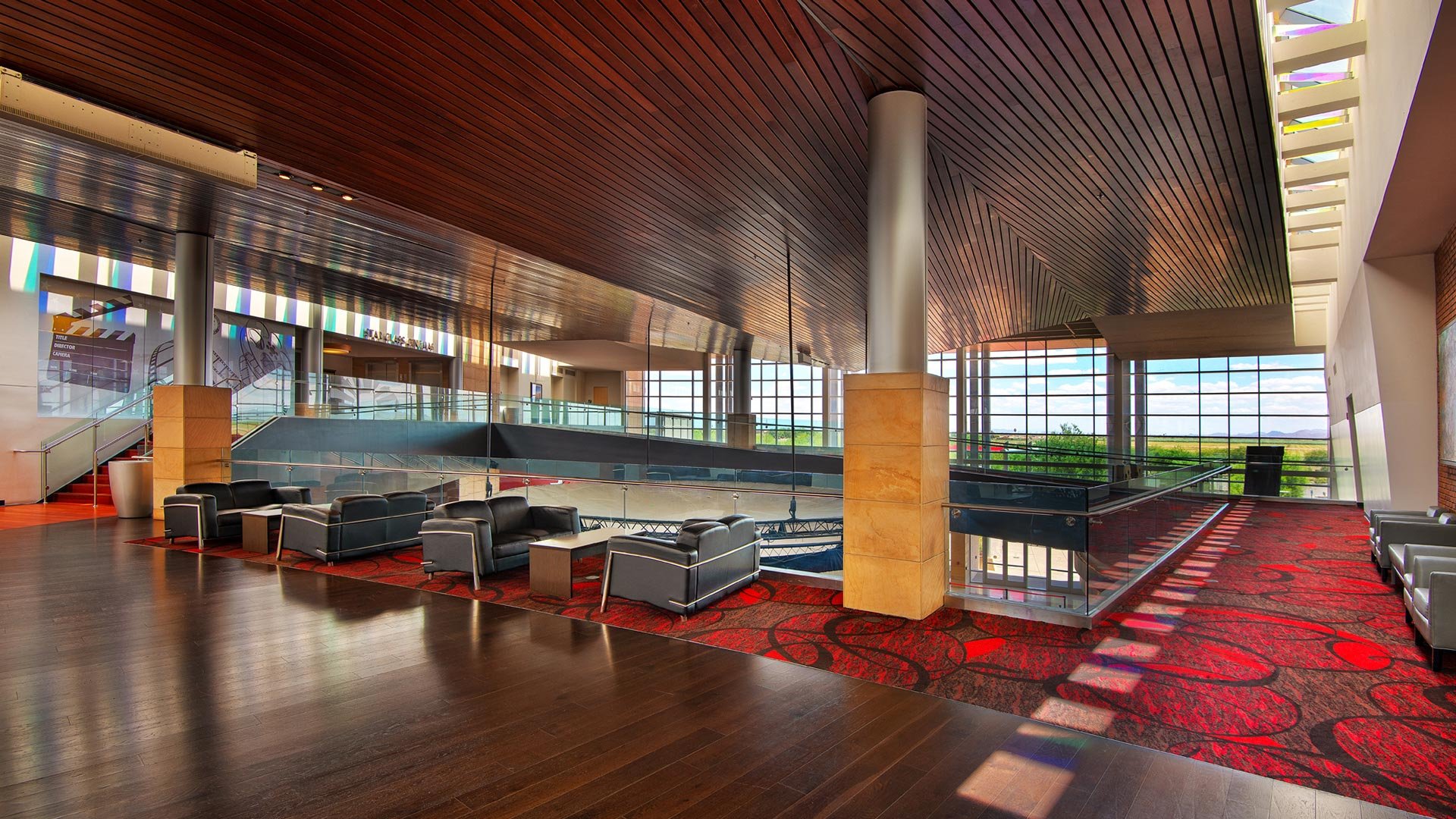 Modern, spacious interior with black leather chairs and small tables on a red patterned carpet, large windows with a view of green fields, wooden ceiling, glass railing, and natural lighting.