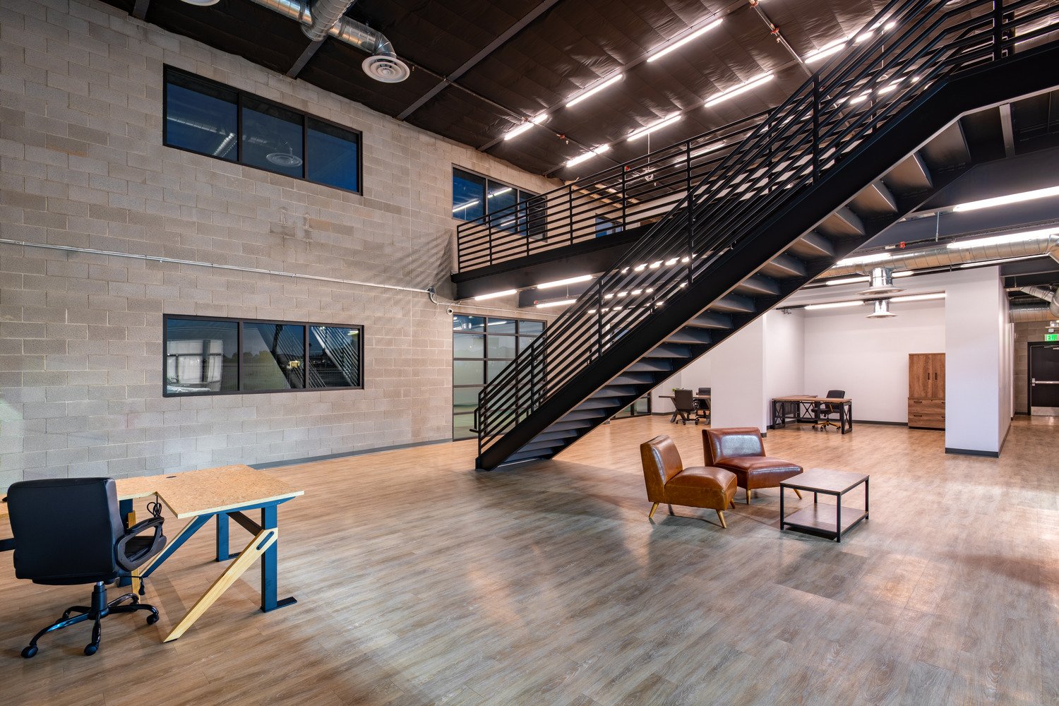 Modern open office space with a staircase, leather chairs, desk, and large windows.