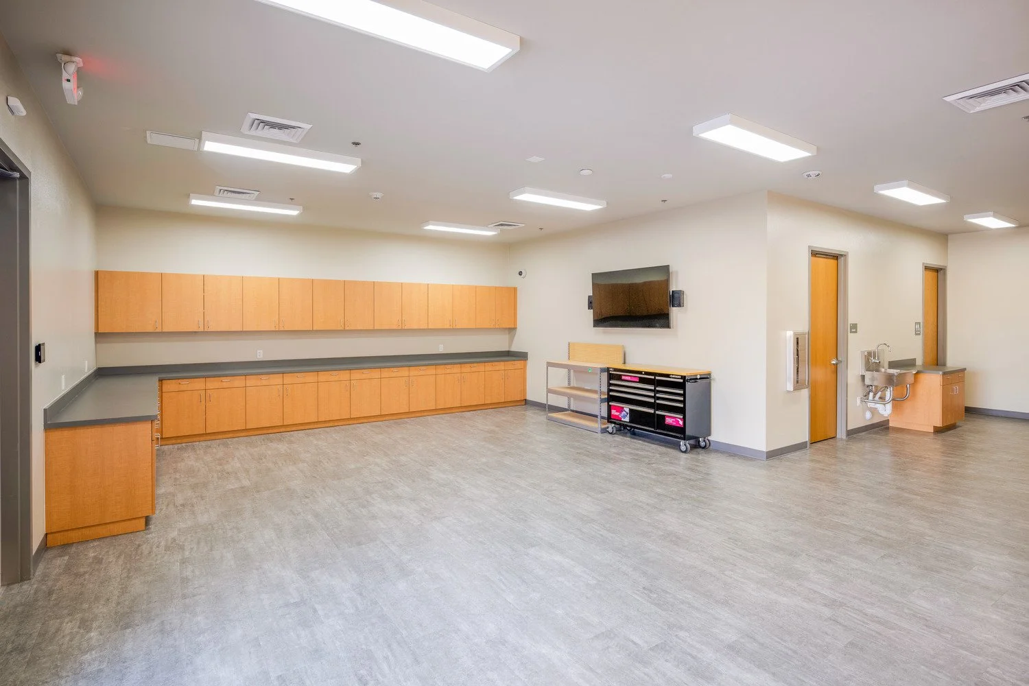 Empty hospital or clinic room with wooden cabinets, a wall-mounted monitor, a utility cart, a sink, and fluorescent lighting.