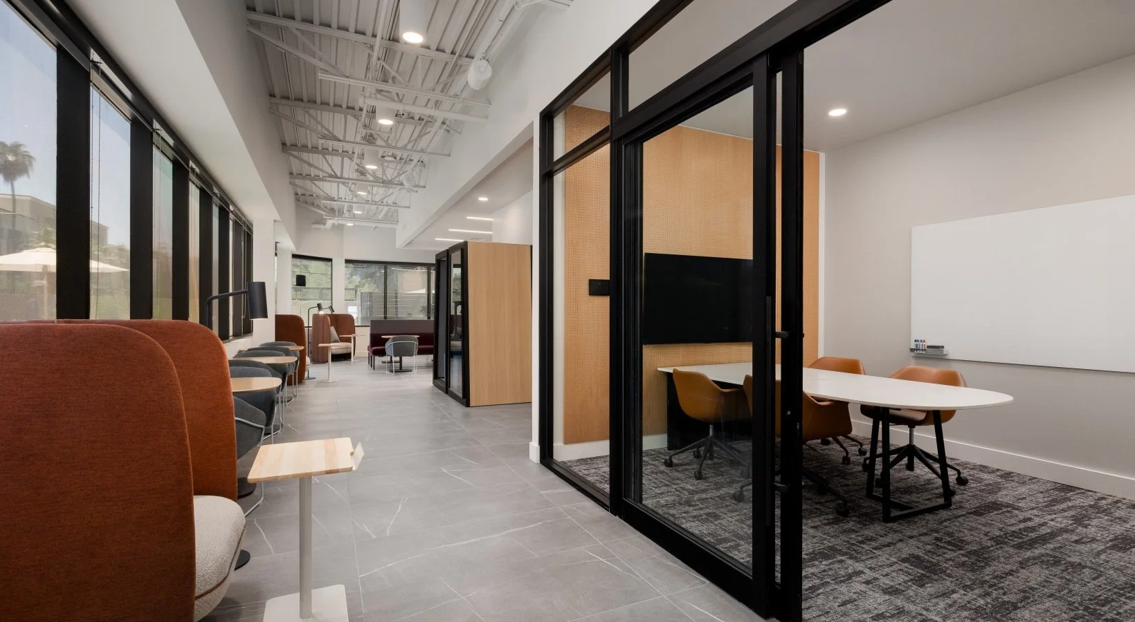 Modern office lounge area with large windows, orange and gray chairs, a small table, and a glass-enclosed meeting room with a whiteboard and a table with four chairs.