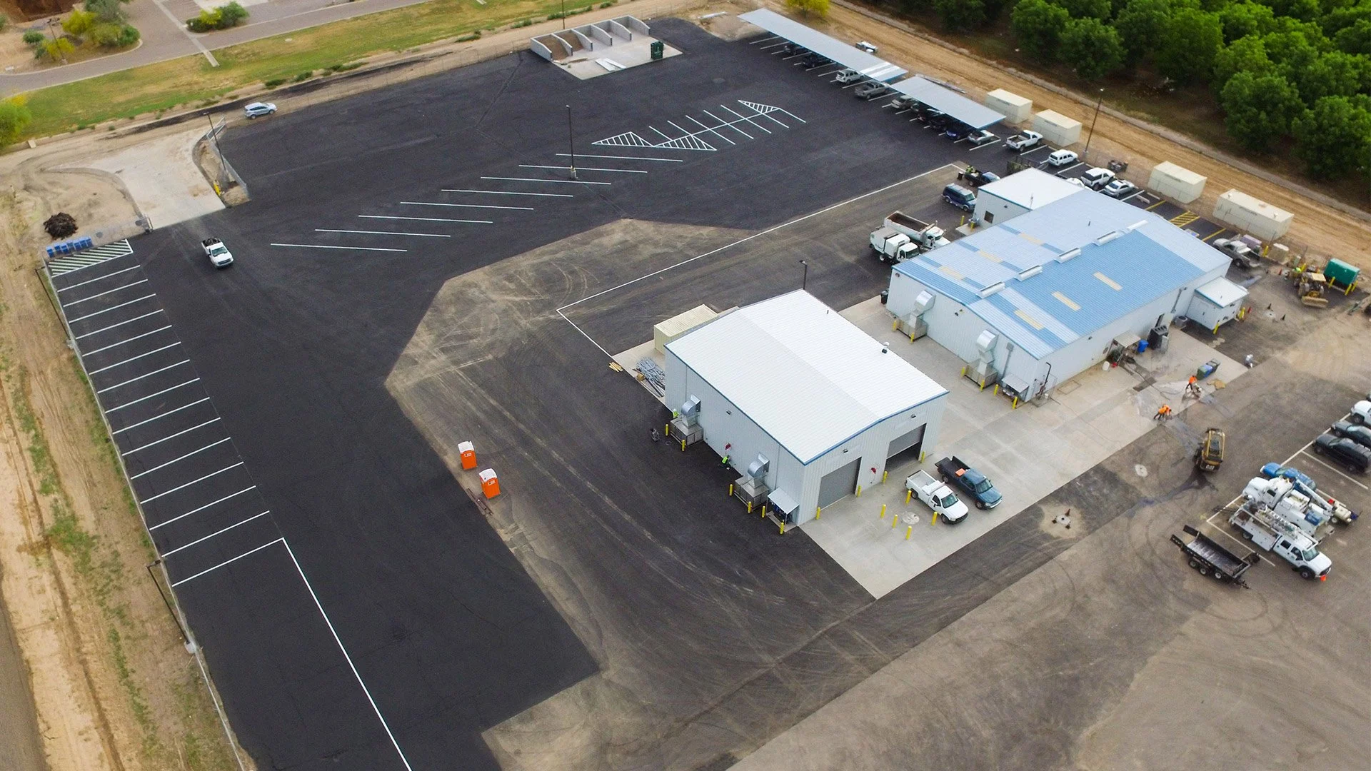 An aerial view of a newly paved parking lot with marked parking spaces, two industrial buildings, and several vehicles parked around the premises.