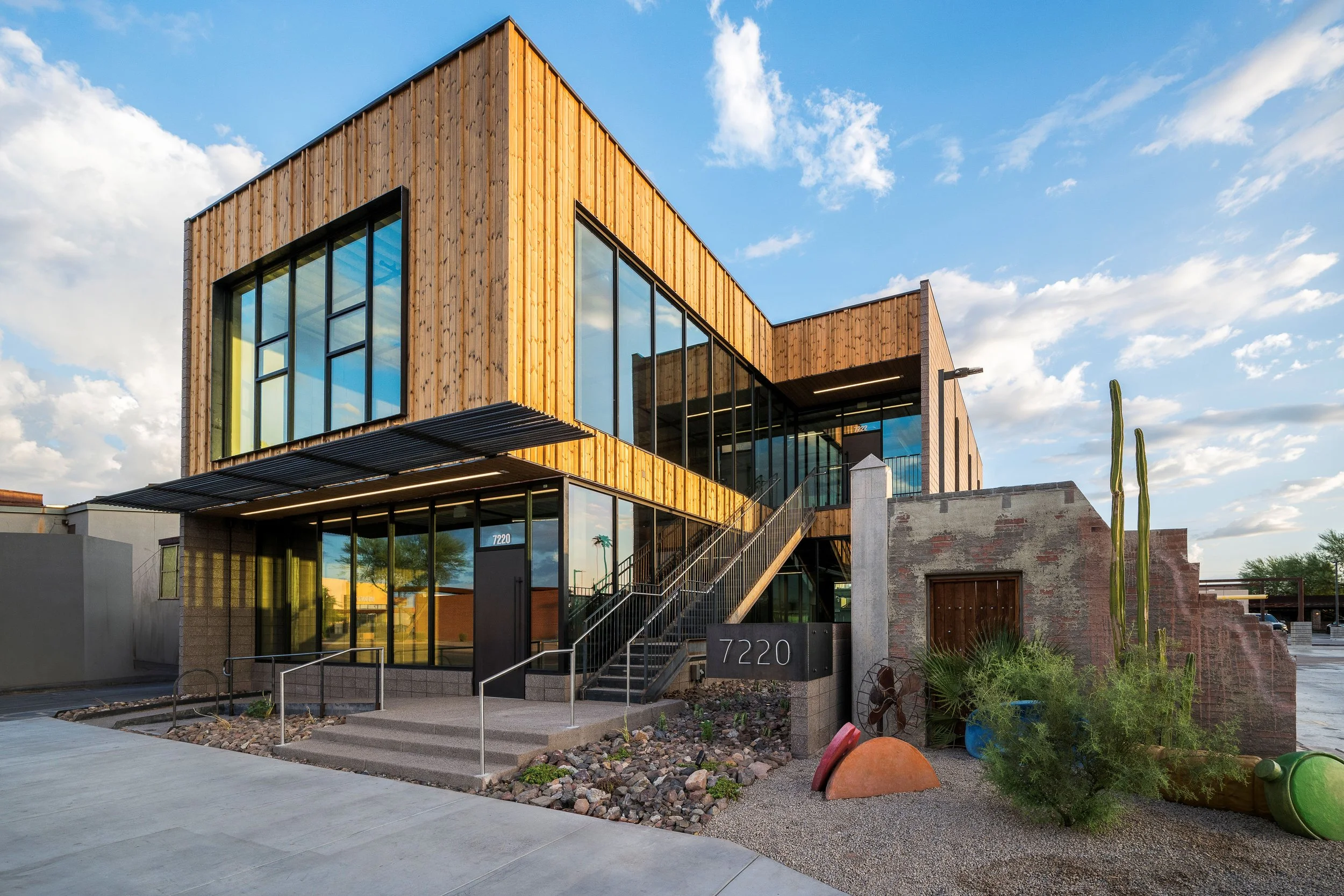 Modern two-story building with wooden siding, large glass windows, stairs on the front, cactus plants, and a blue sky with clouds.