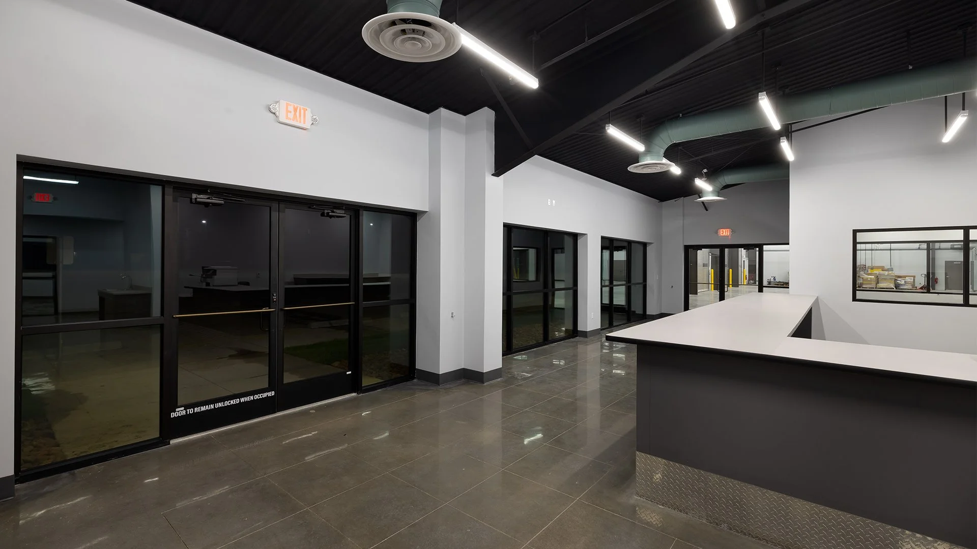 Empty modern lobby or reception area with black-framed glass doors and windows, white walls, polished concrete floor, and ceiling with exposed ductwork and linear LED lighting.