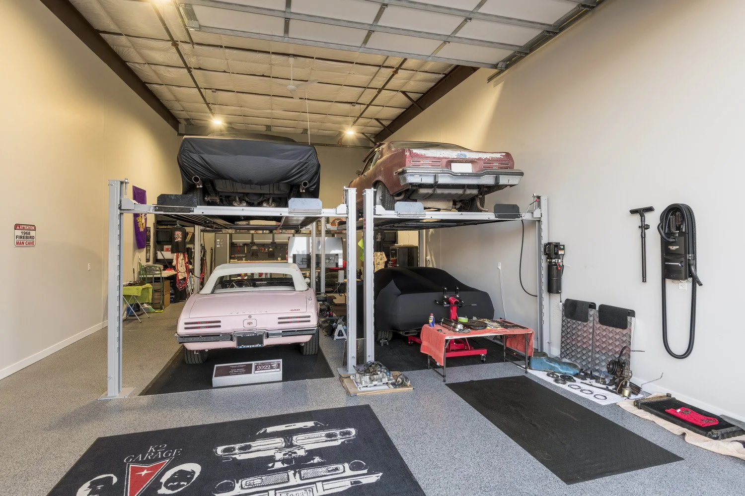 A garage with a two-level car lift holding a black car covered with a cover on one side and an old, dusty red car on the other side. There is a vintage pink car parked below the lift. The garage has various tools, car parts, and equipment, including 