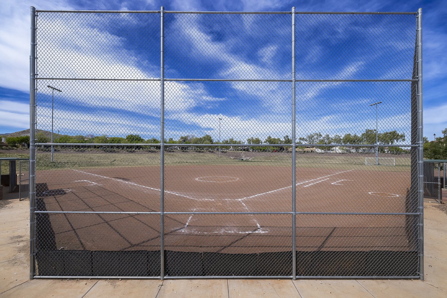 Empty baseball or softball field with a chain-link backstop, dirt infield, and grassy outfield under a partly cloudy sky.