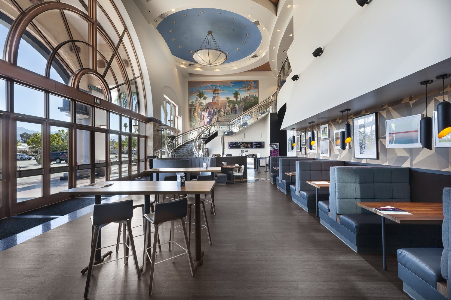 Interior of a modern restaurant or cafe with large front windows, high ceilings, a mural on the back wall, and a staircase leading upstairs. There are booths along the right side and tables with chairs on the left. The decor includes framed pictures 