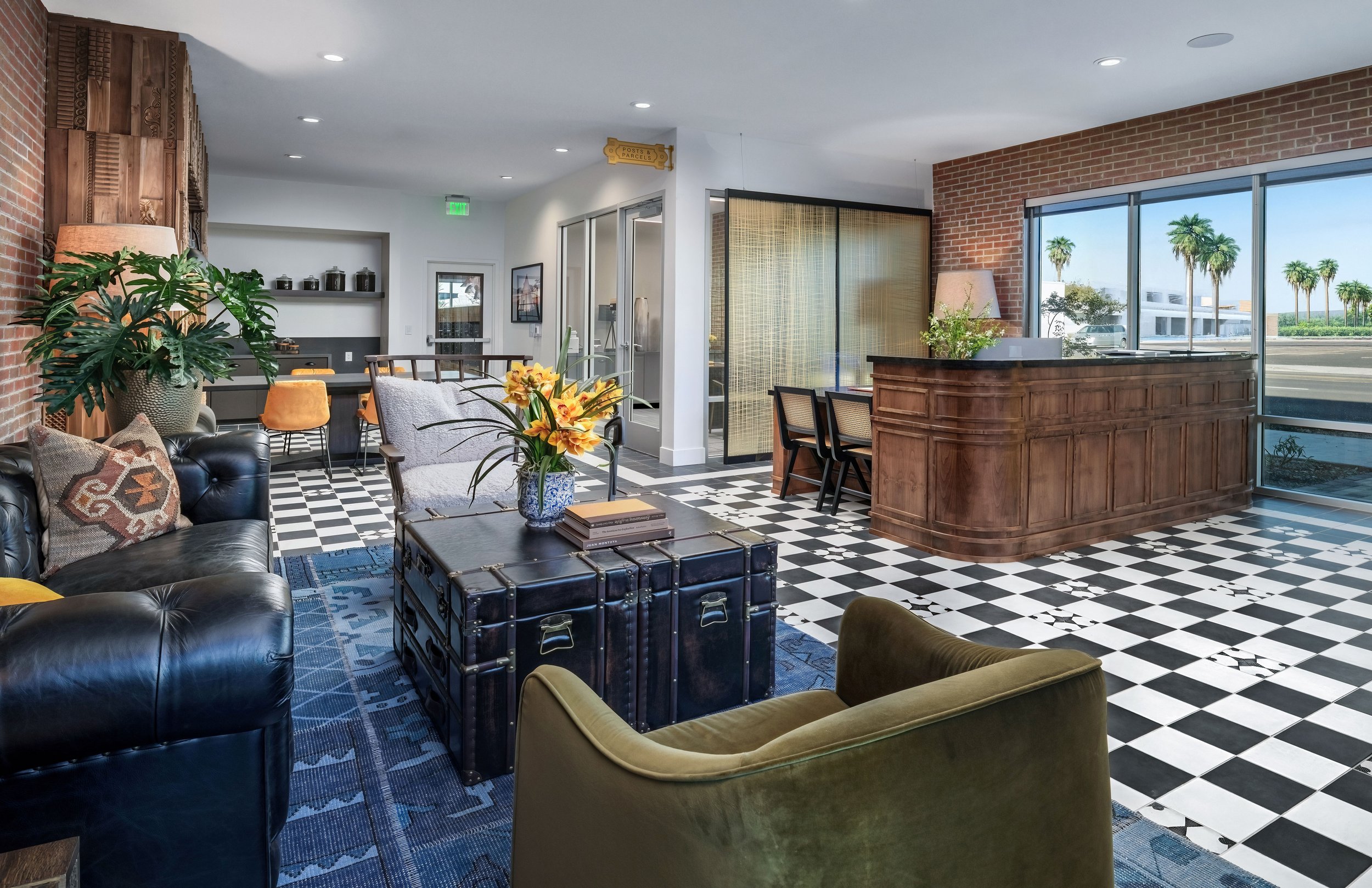 Interior of a modern lounge with a black leather sofa, green armchair, coffee table with flowers, checkered floor, wooden reception desk, large windows with palm trees outside, and a small kitchen area in the background.