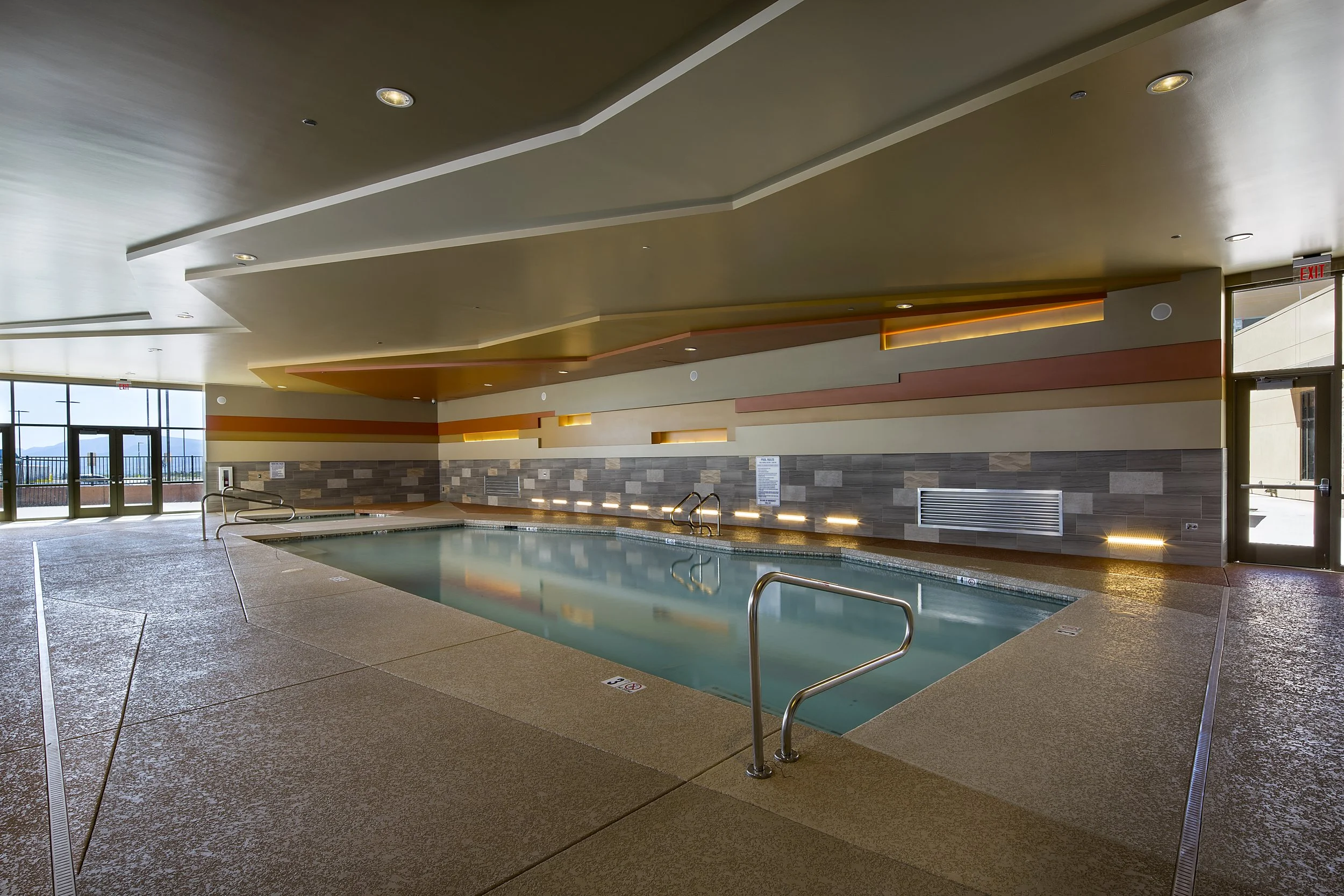 Indoor swimming pool with stainless steel handrails, tiled wall, and large glass doors with a view outside.