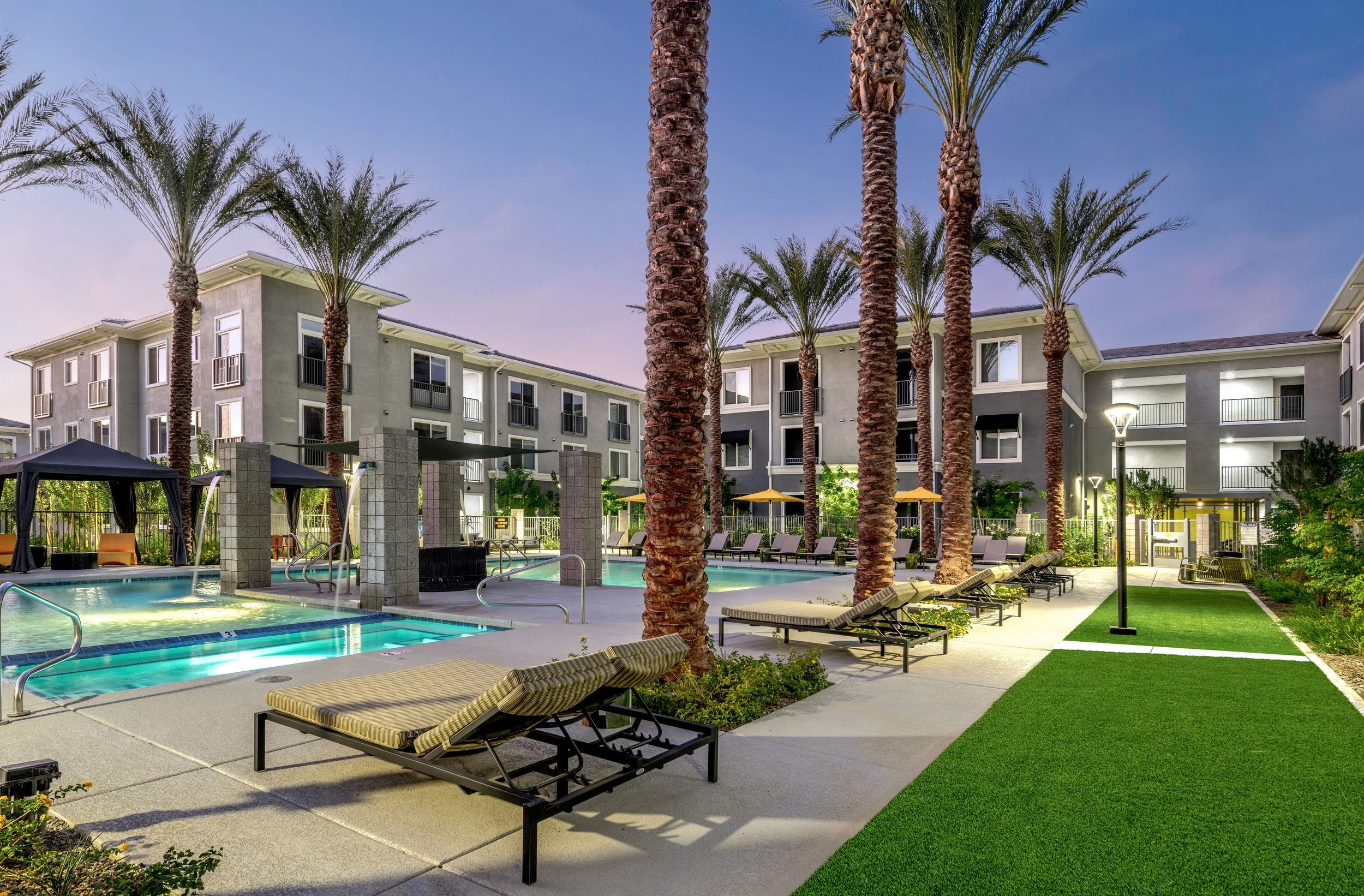 Stylish outdoor pool area in an apartment complex with lounge chairs, palm trees, and apartment buildings in the background during dusk.