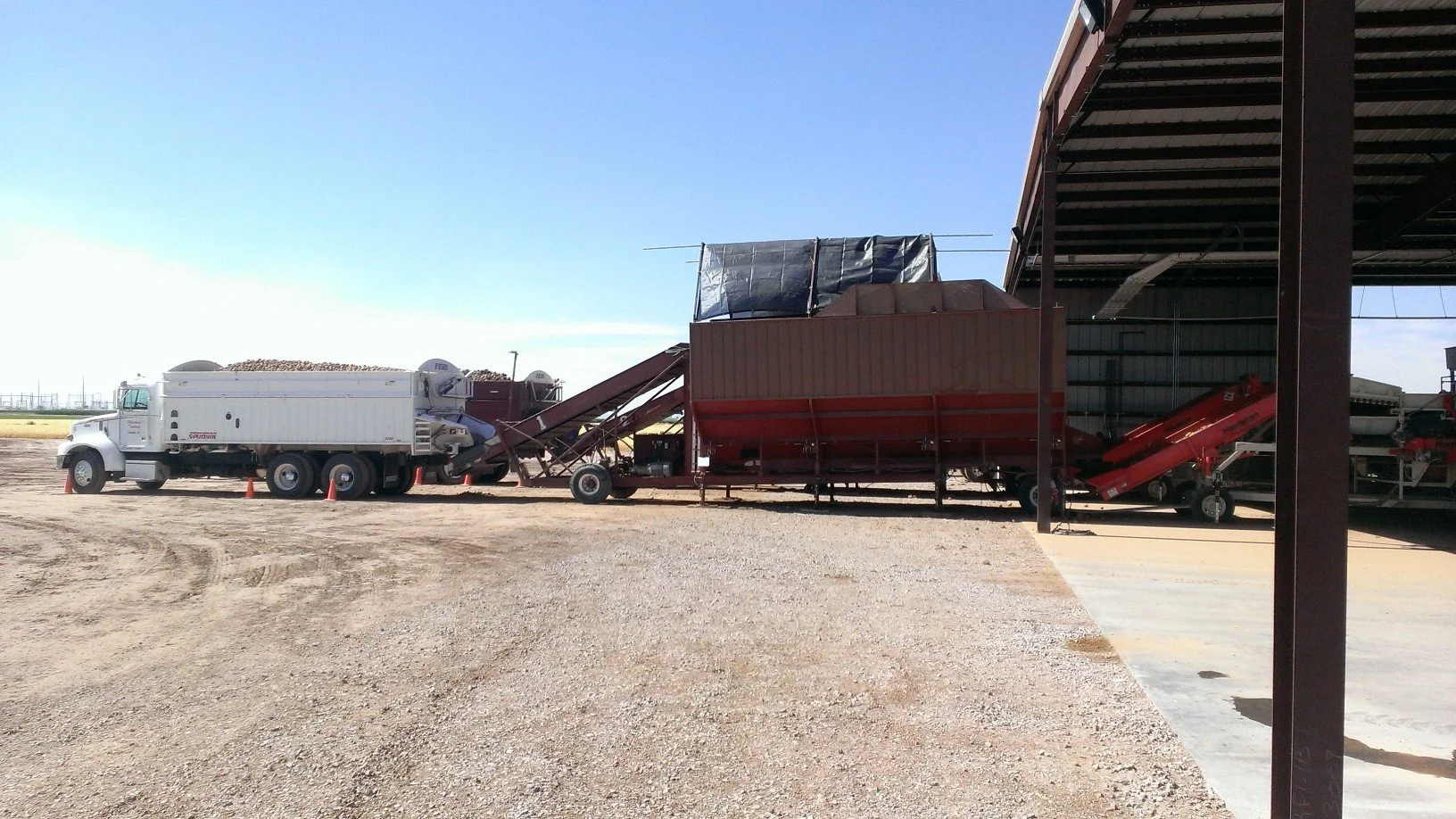 Agricultural equipment and trucks outside a large storage shed on a farm or industrial site, with clear sky and gravel ground.