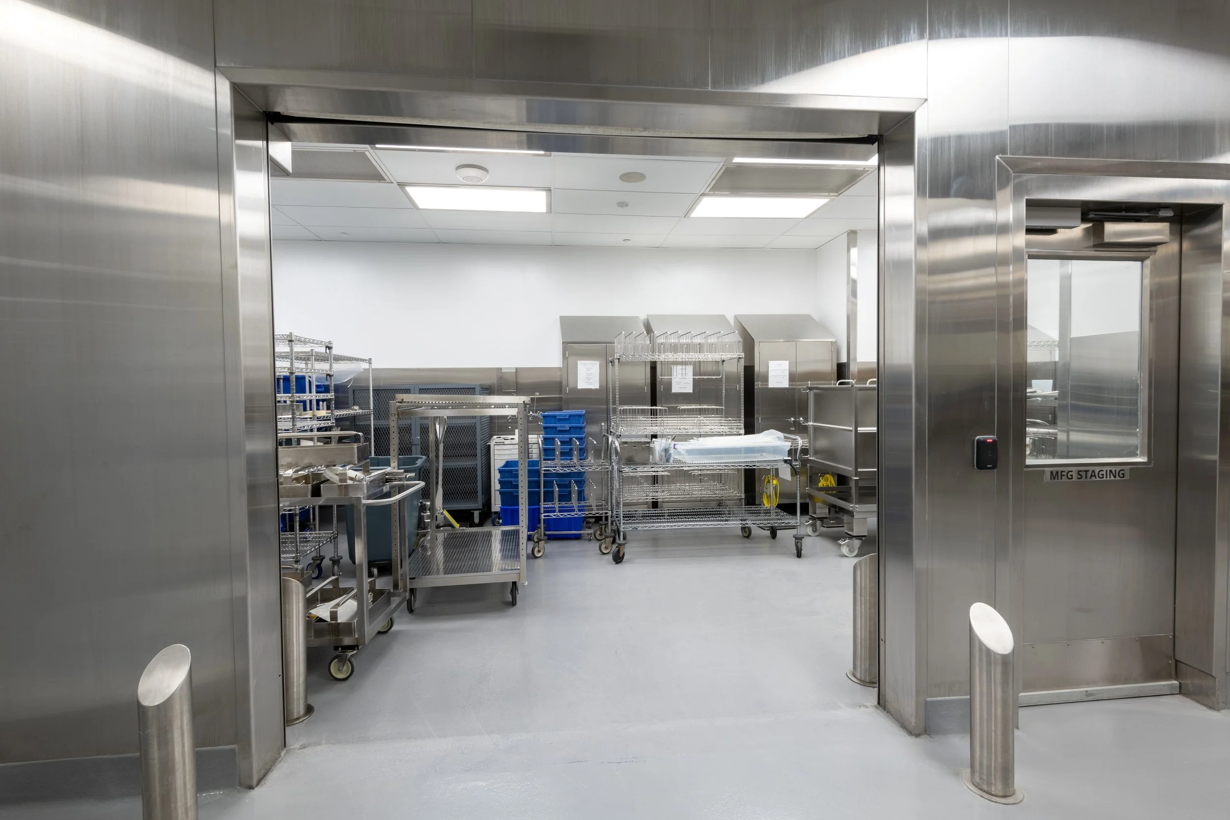 Empty sterile medical storage room with stainless steel shelves and carts, viewed through an open doorway.
