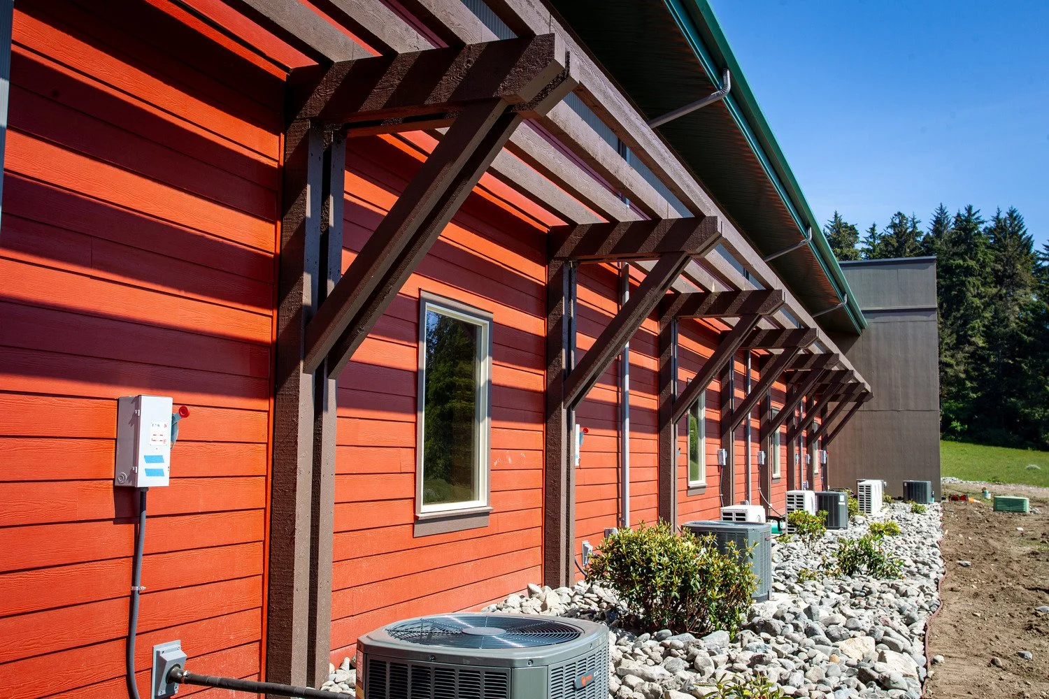 Side view of a red building with multiple air conditioning units on the ground outside, several windows, and a series of wooden awnings above each window, with a green roof and lush trees in the background.