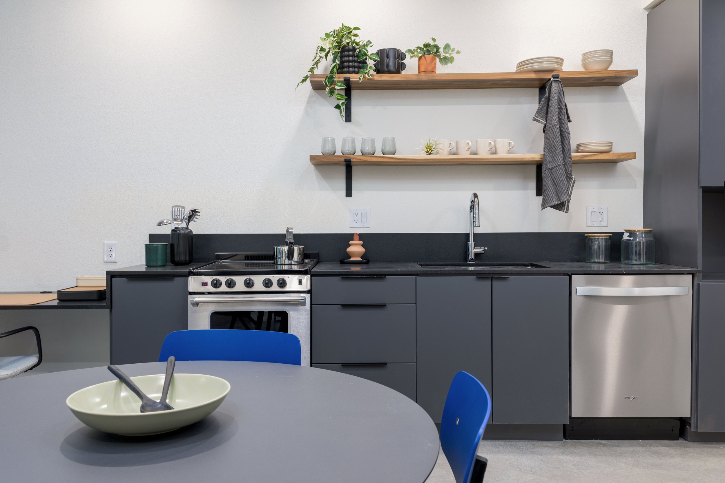 Modern kitchen with gray cabinets, black countertop, and open wooden shelves holding dishes, cups, and plants. A round table with blue chairs and a bowl with utensils is in the foreground.