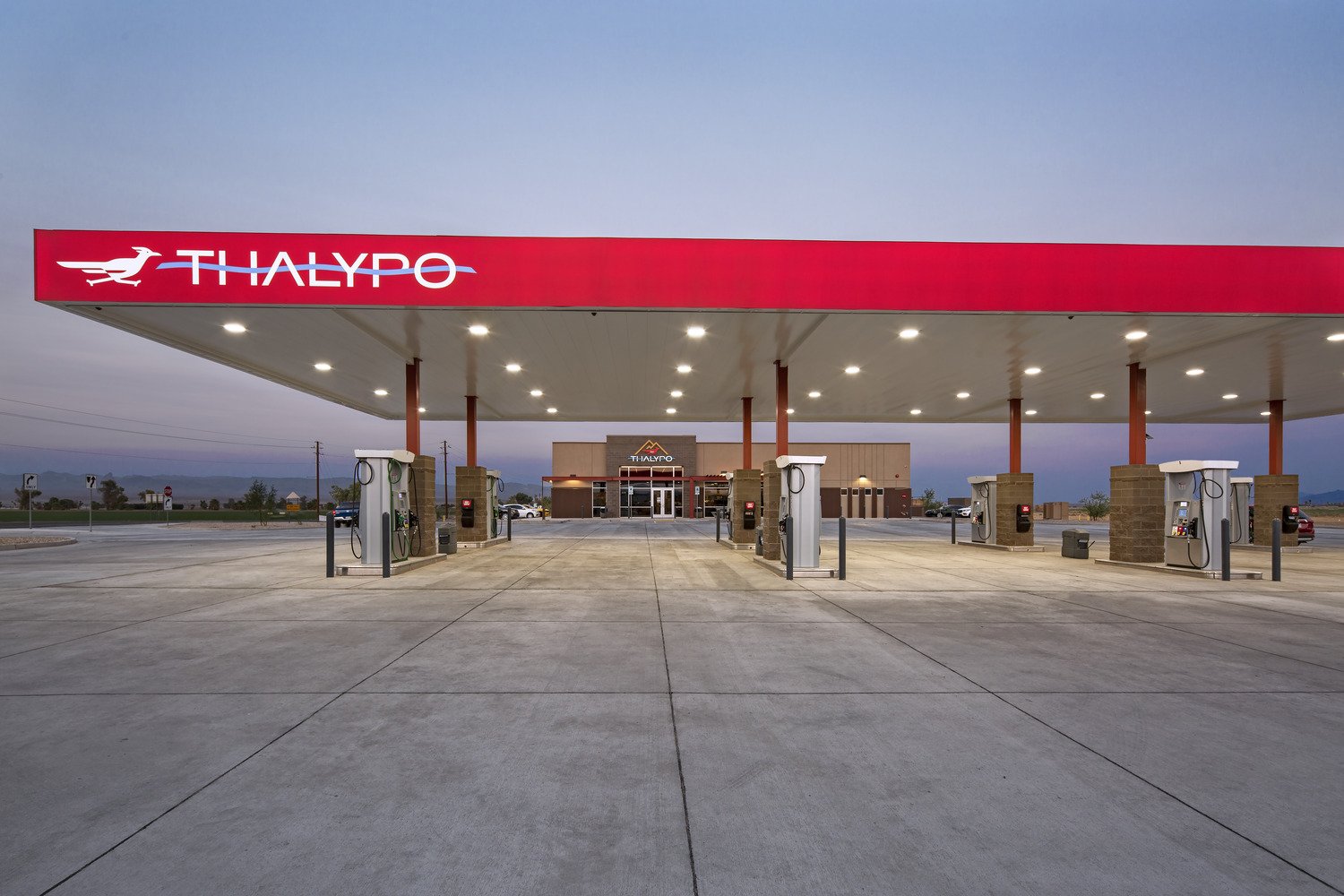 Empty Thalpyo gas station with multiple fuel pumps under a large red canopy, with a store or building in the background, during dusk or dawn with a cloudy sky.
