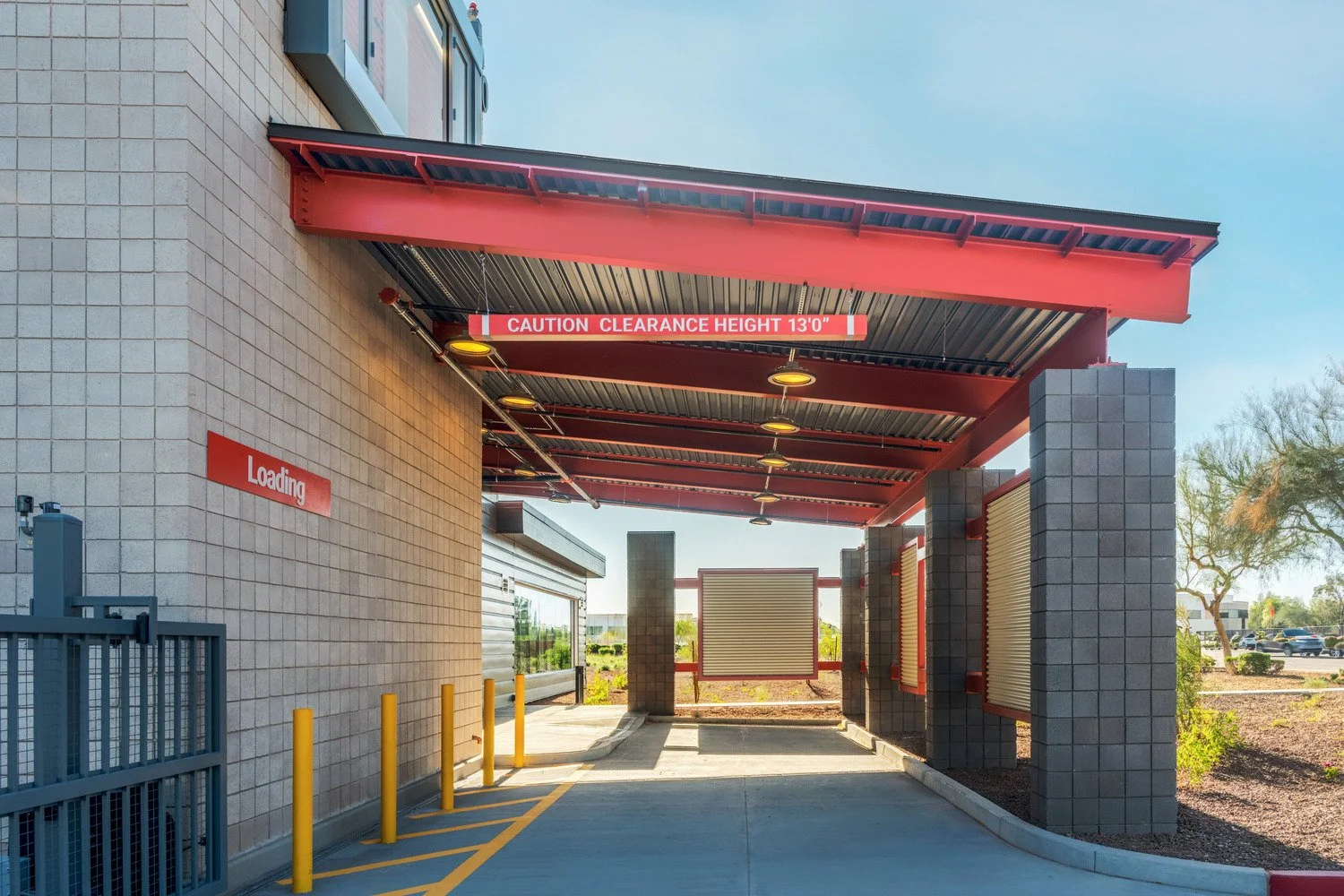 A drive-thru loading area with a sign saying 'Loading' on the left wall and a red clearance height sign overhead reading 'CAUTION CLEARANCE HEIGHT 13'0"'. The area has yellow bollards, gray brick walls, and a red metal roof structure with multiple li