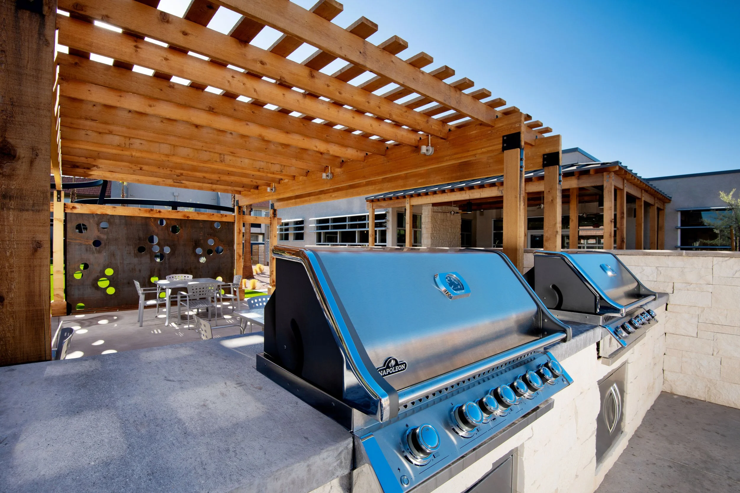 Outdoor kitchen area with stainless steel grills, a stone countertop, a wooden pergola overhead, and a patio with tables and chairs in a sunny setting.