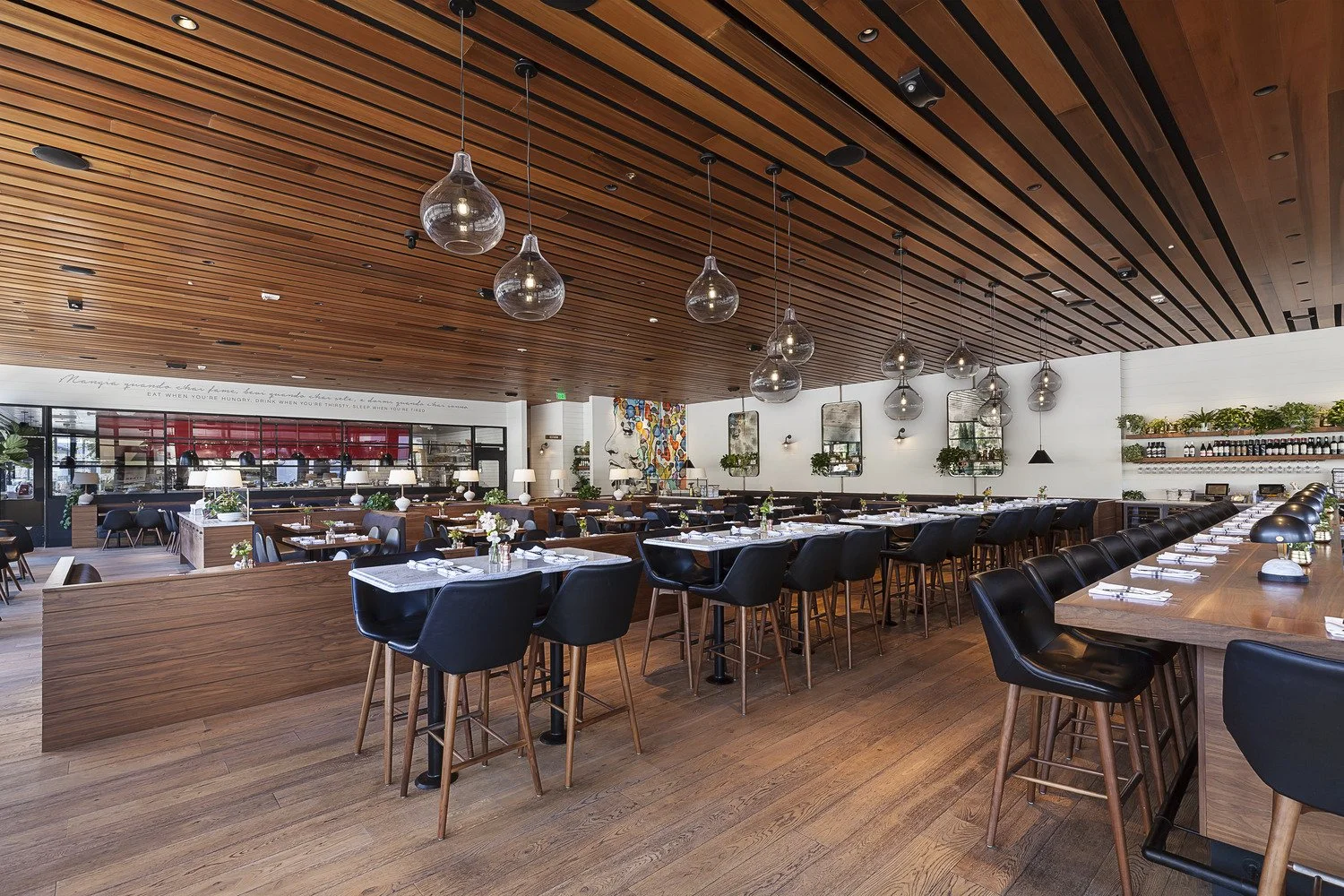 Interior of a modern restaurant with wooden ceiling and flooring, black chairs, long wooden tables set with white napkins, small vases with flowers, decorative wall shelves with plants, hanging glass pendant lights, and a colorful abstract painting o