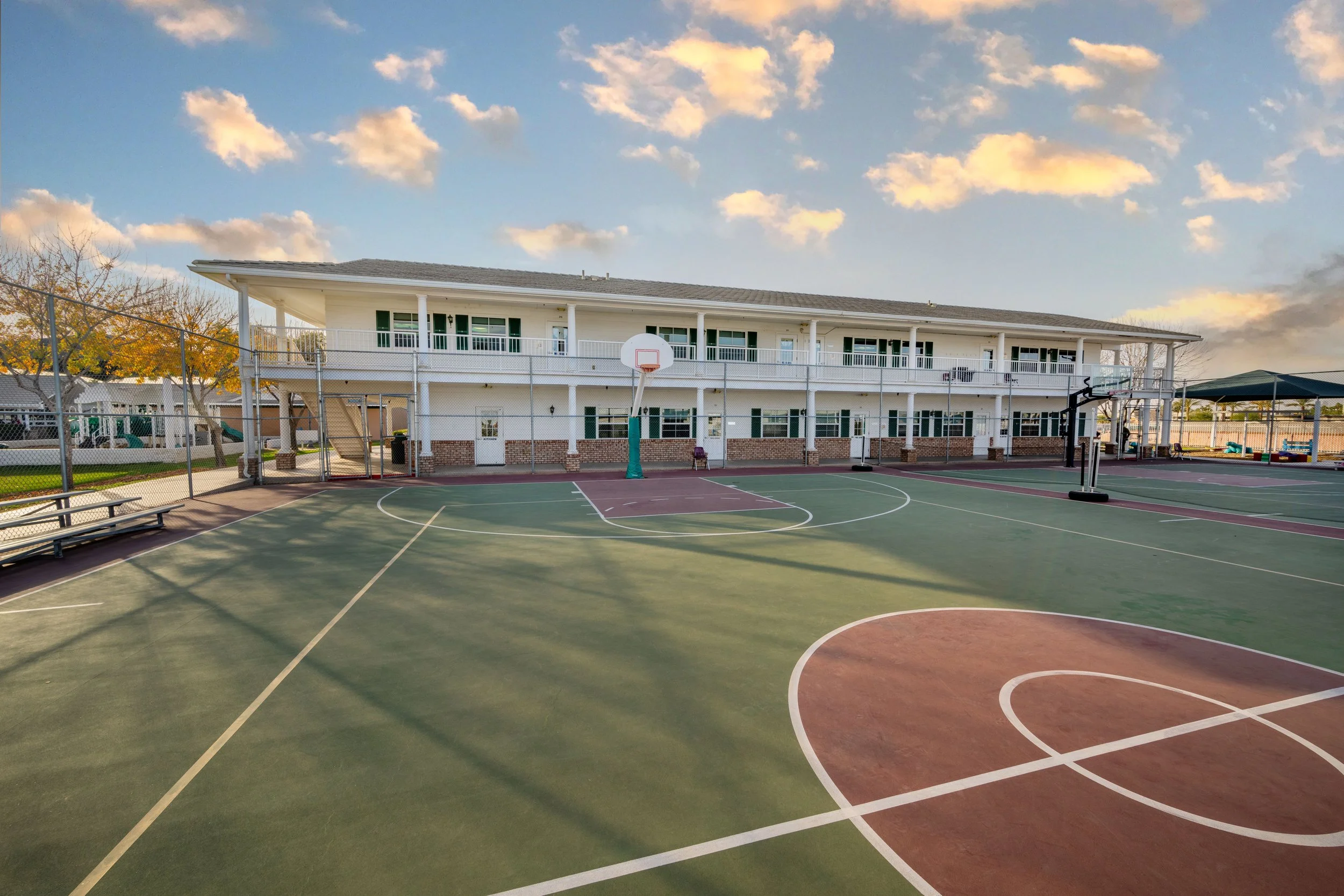 Empty outdoor basketball court with a building in the background on a sunny day with some clouds.